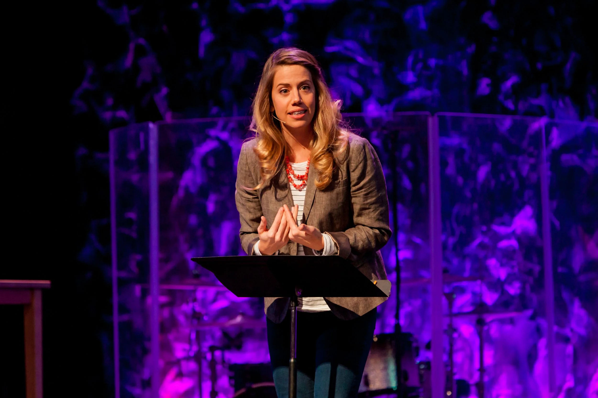 A woman speaking at a podium during a presentation or event, with a dark background illuminated by purple lighting, wearing a gray blazer and a red necklace.
