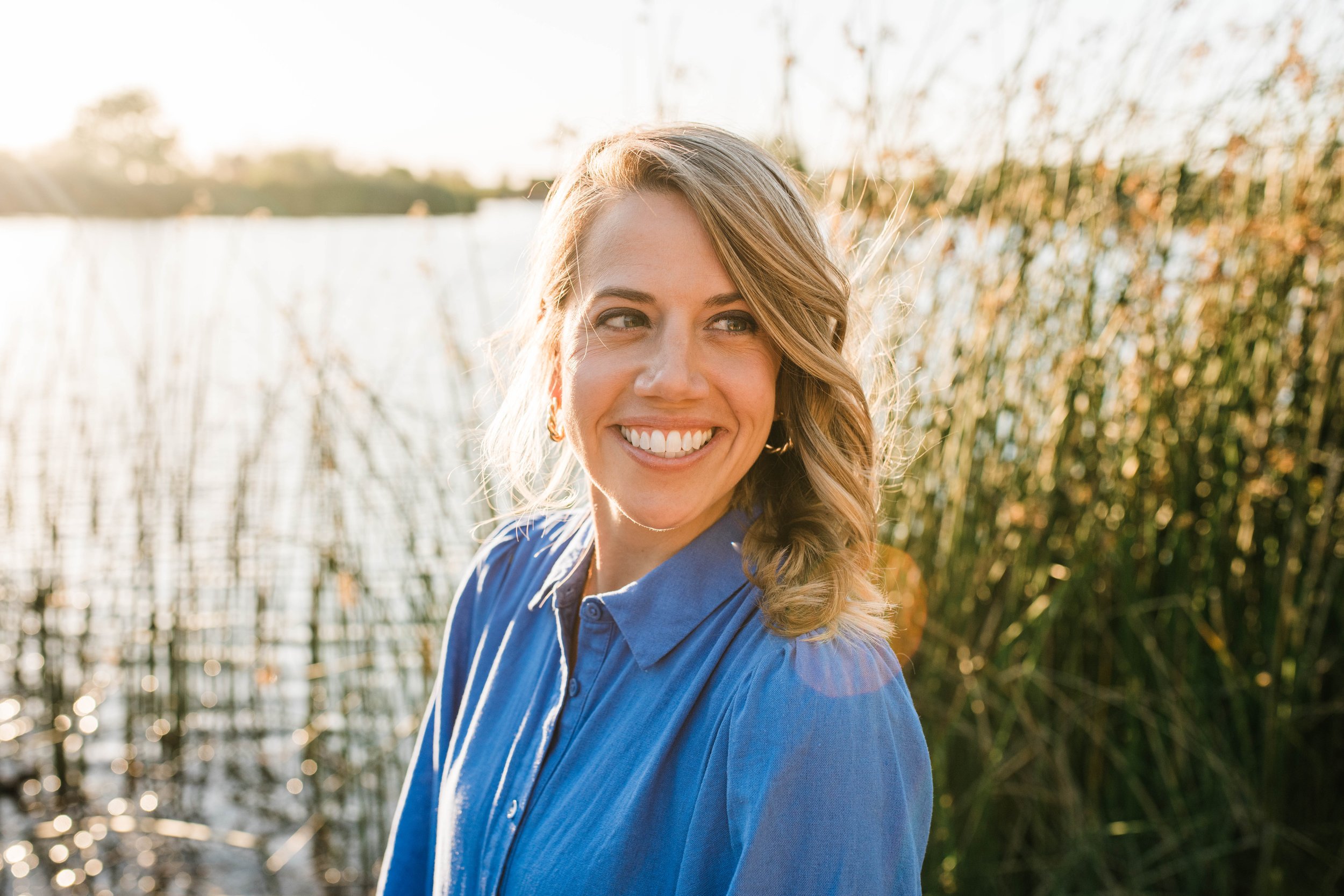 A woman smiling outdoors with sunlight reflecting on water and tall grass or reeds in the background.