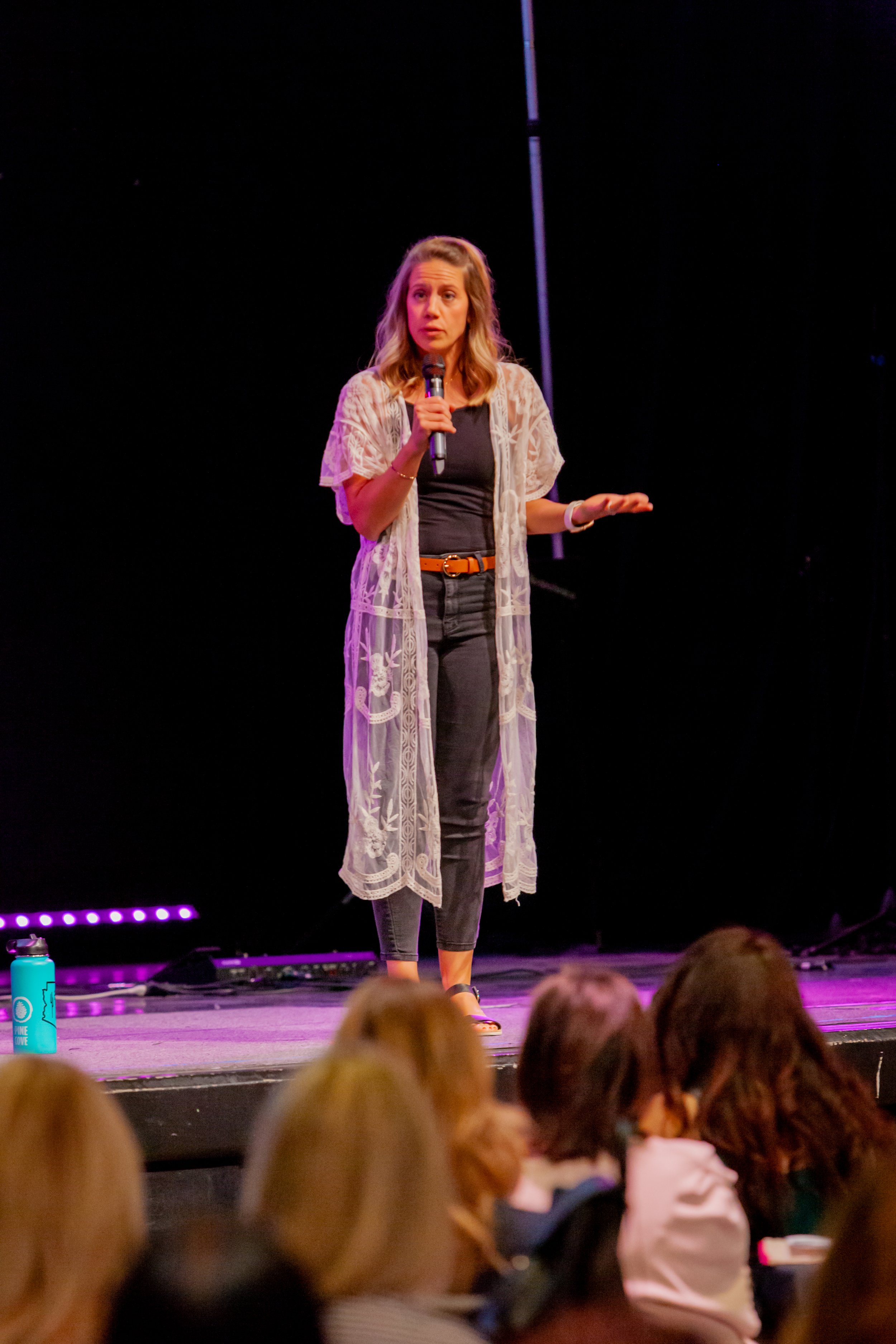 A woman standing on stage speaking into a microphone, wearing a dark top, gray pants, and a lacy, long, white cardigan, with an audience in the foreground.