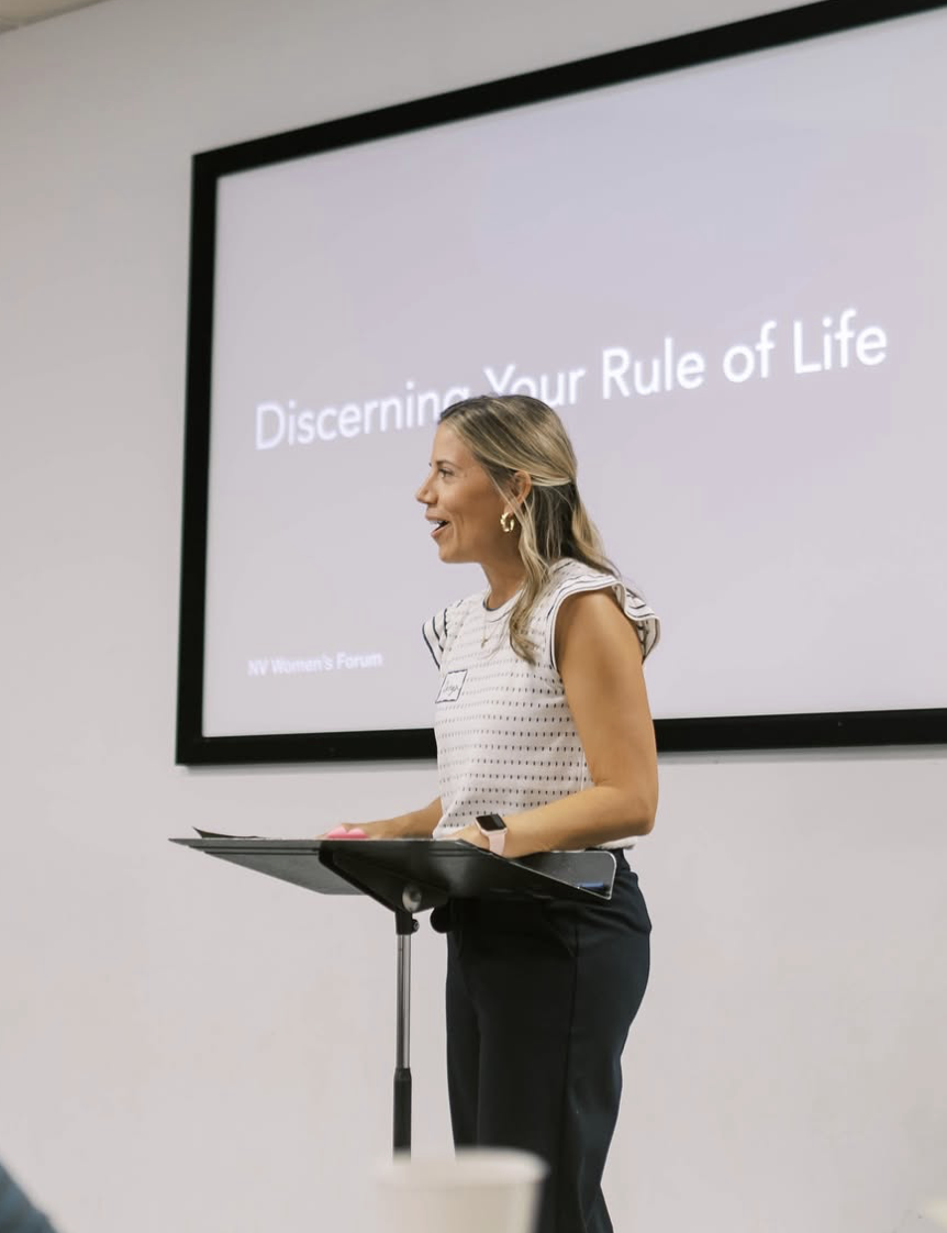 A woman presenting at a seminar or conference, standing at a podium with a large screen behind her displaying the title "Discerning Your Rule of Life."