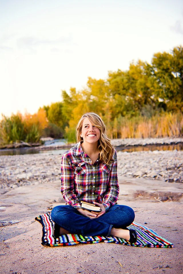 A young woman sitting cross-legged on a colorful striped blanket outdoors near a river, holding books, smiling, with trees and a bright sky in the background.