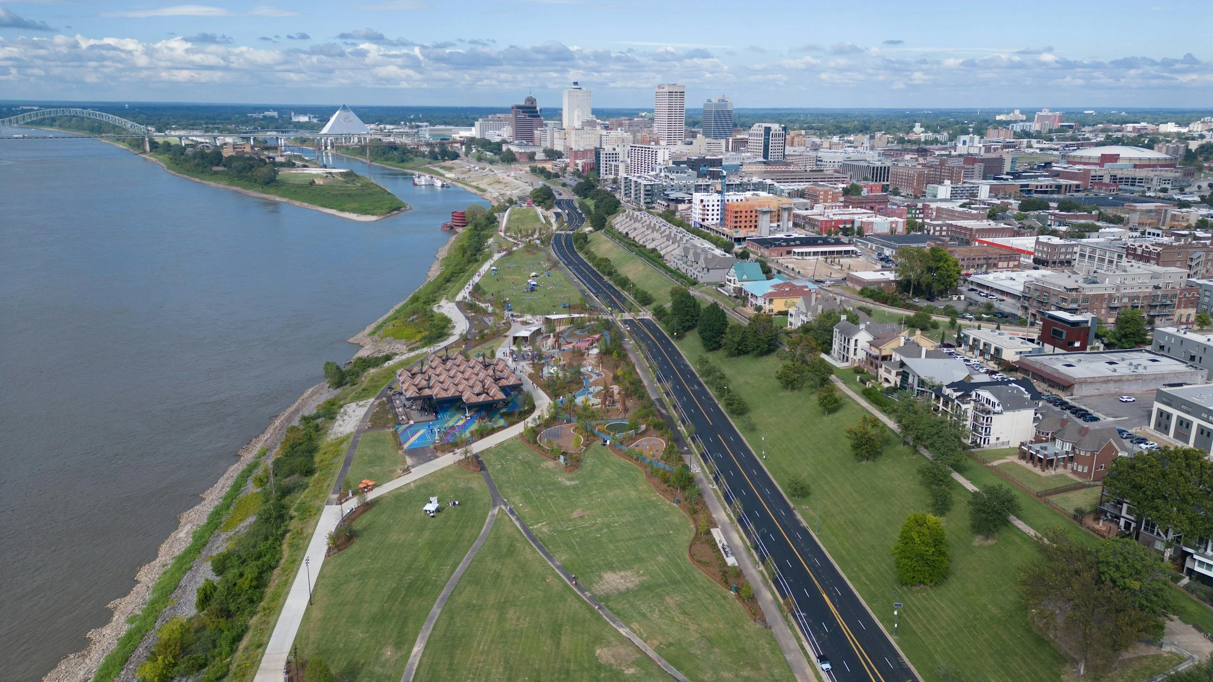 Aerial view of a cityscape along a river, with green parks, walking paths, playgrounds, and a downtown area with tall buildings.