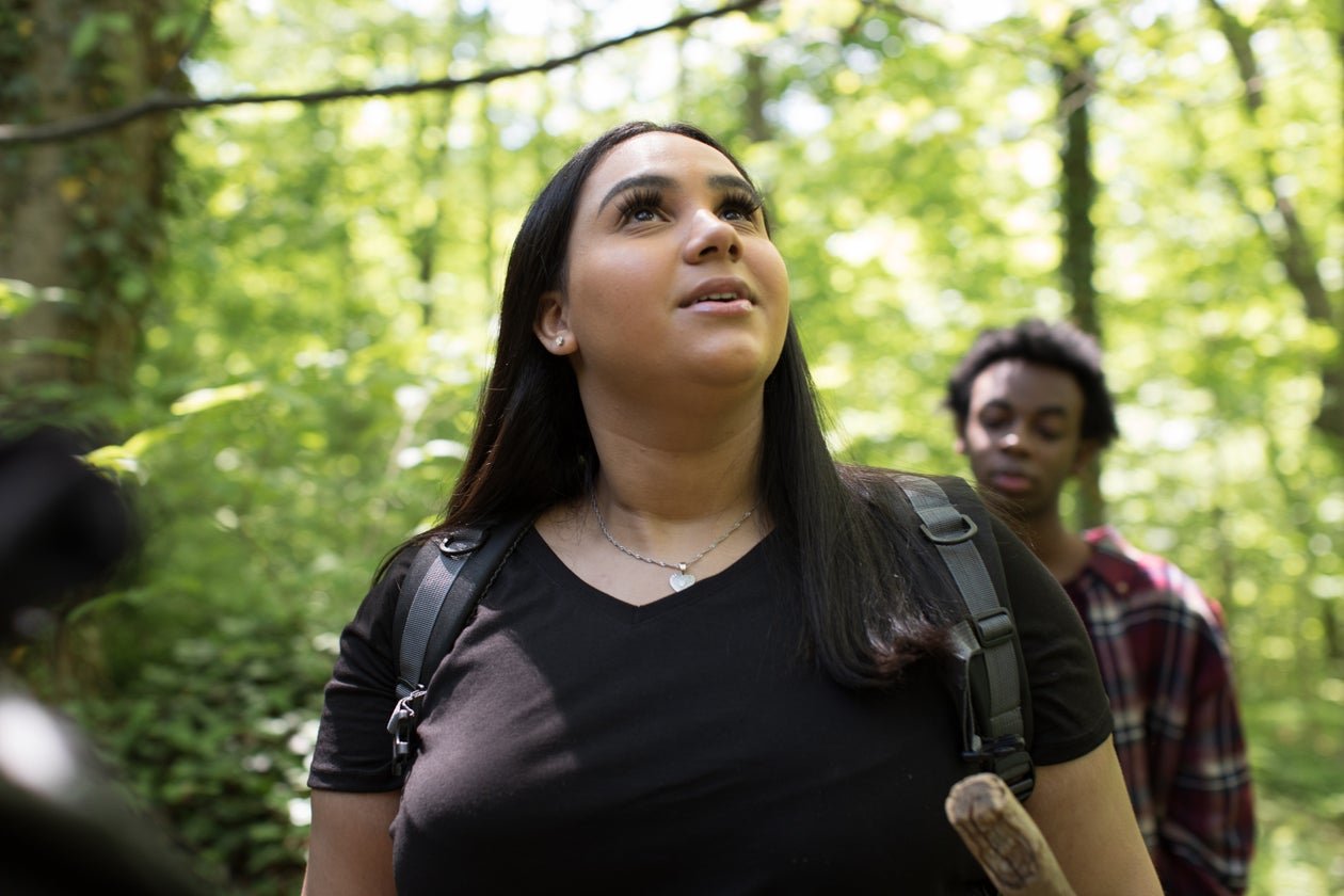 Two women hiking through a green, leafy forest, with one woman in front looking upward and the other woman slightly behind, both carrying backpacks.