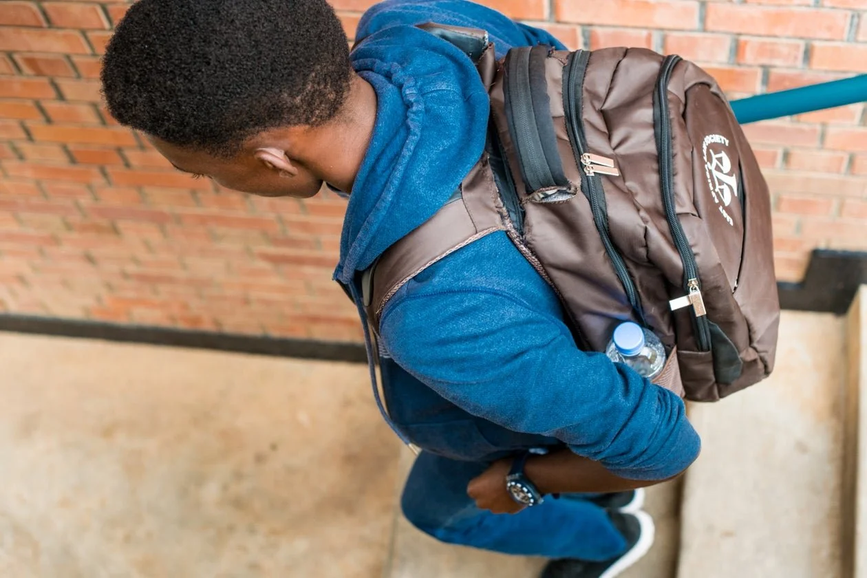 Top-down view of a young man wearing a blue hoodie, jeans, and sneakers, carrying a brown backpack with a water bottle, standing with arms crossed against a brick wall background.