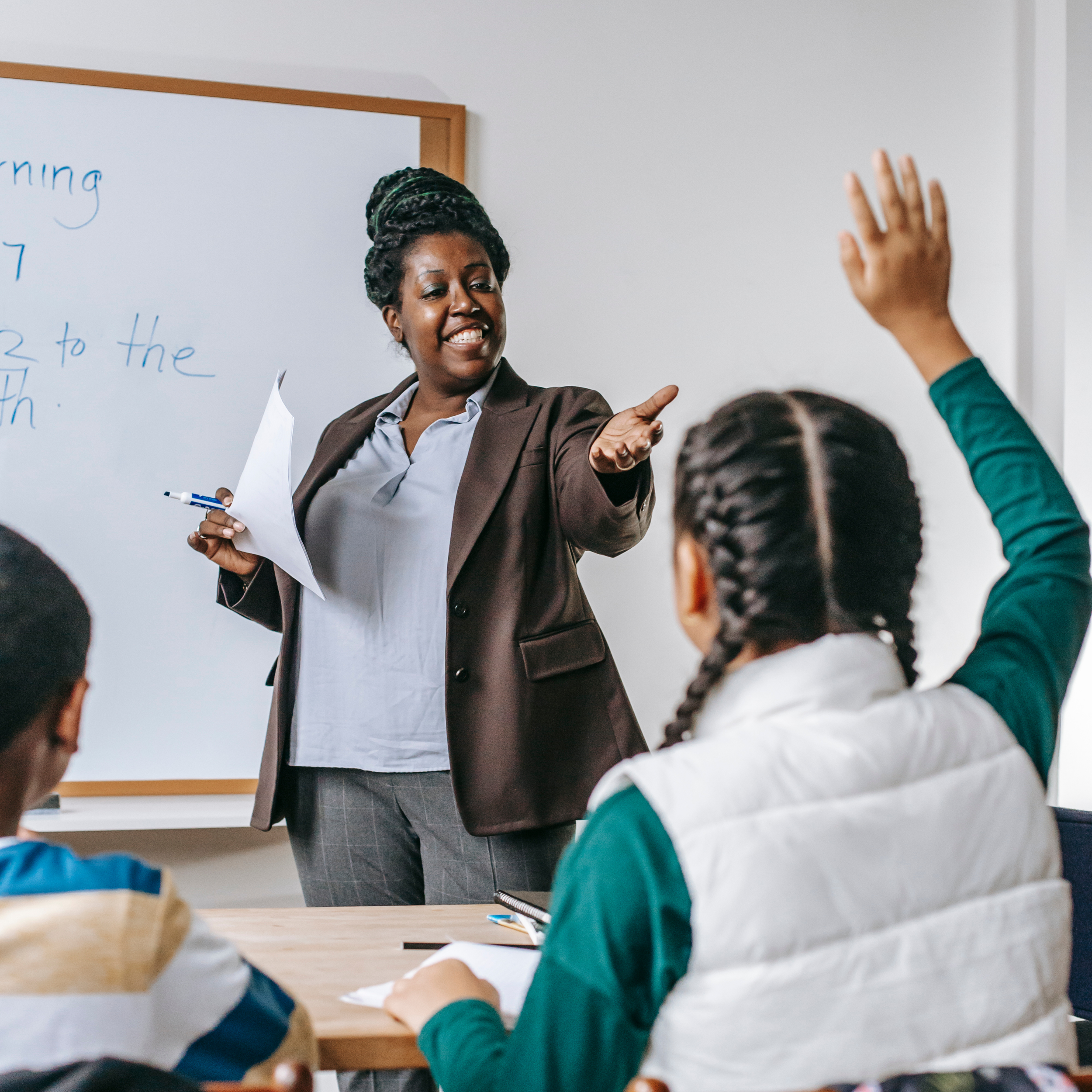 Teacher standing in front of a whiteboard, gesturing towards a student who has raised her hand during a classroom lesson.