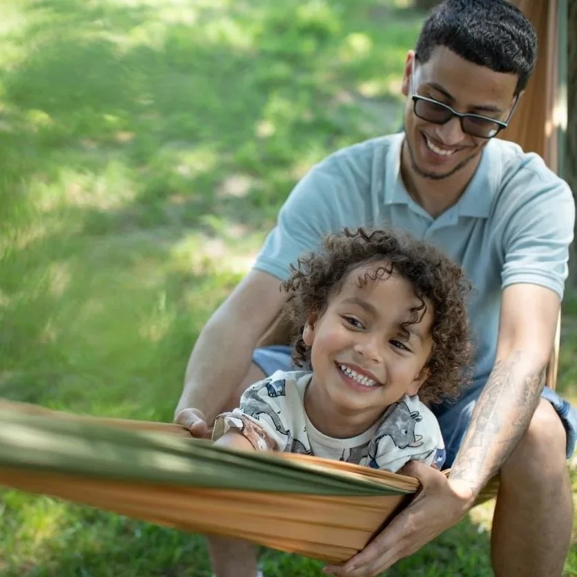 A young boy and an adult man playing outside in a swing or hammock, smiling and enjoying a sunny day with green trees in the background.