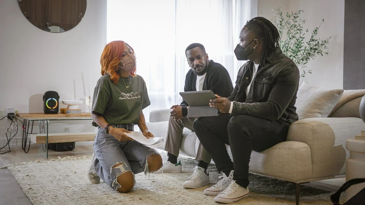 Three people in a living room having a discussion, with two people sitting on a sofa and one kneeling with documents, all wearing masks.
