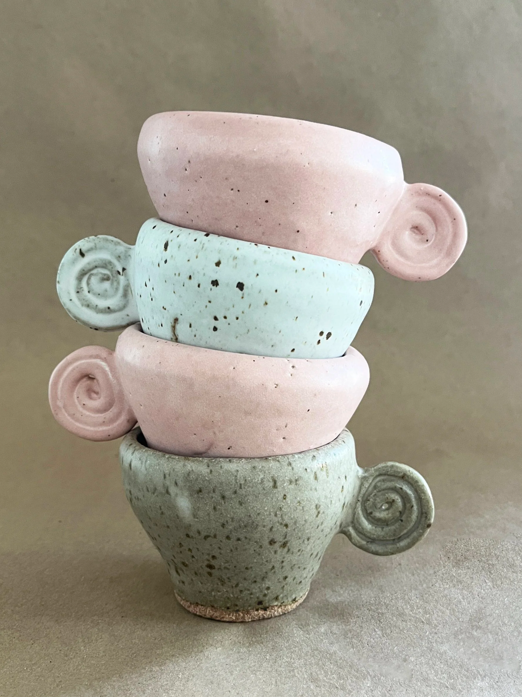 A stack of five ceramic cups with spiral handles, in pastel pink, mint green, and beige colors, with a textured, speckled surface, on a neutral background.