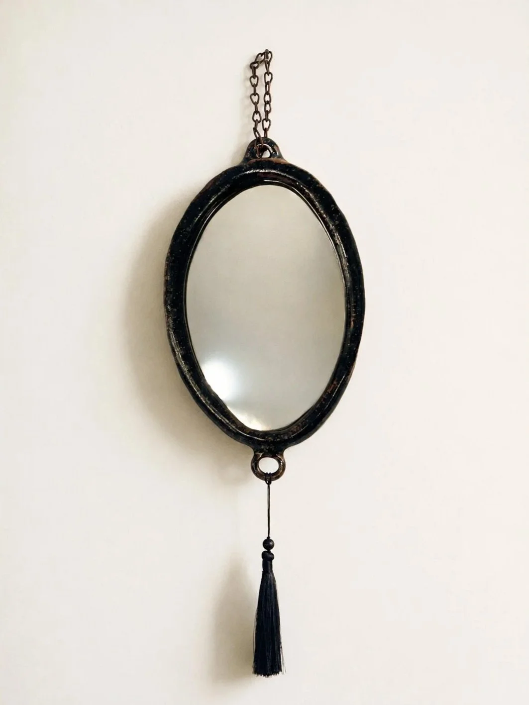 An oval-shaped black vintage mirror with a chain at the top and a tassel hanging from the bottom, mounted on a plain white wall.