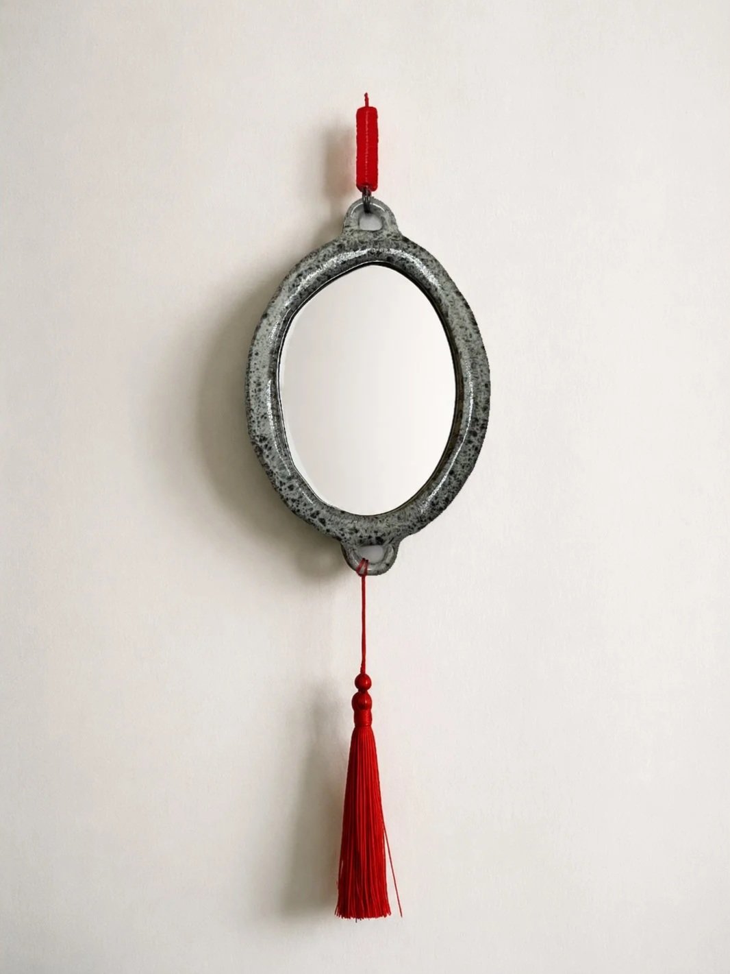 A round mirror with a stone-like frame hanging on a plain white wall, with a red tassel and beads attached to the bottom and a red hook at the top.