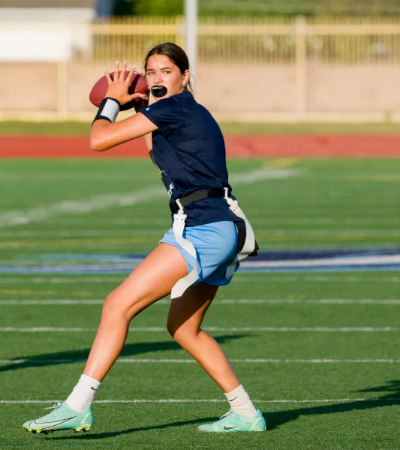 A young female athlete playing football on a field, wearing a navy blue jersey, light blue shorts, and white athletic shoes, holding a football ready to throw.