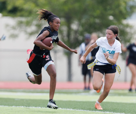 Two young women playing flag football outdoors on a field, one running with a football and the other chasing.