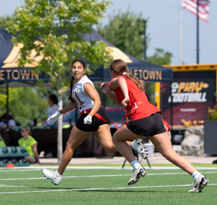 Two female flag football players running on a football field during a game, with a tent, trees, and American flags in the background.