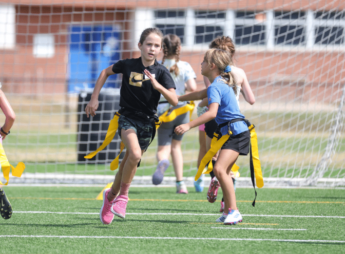 Young girls participating in a flag football game on a grassy field, with a goal net and a school building in the background.