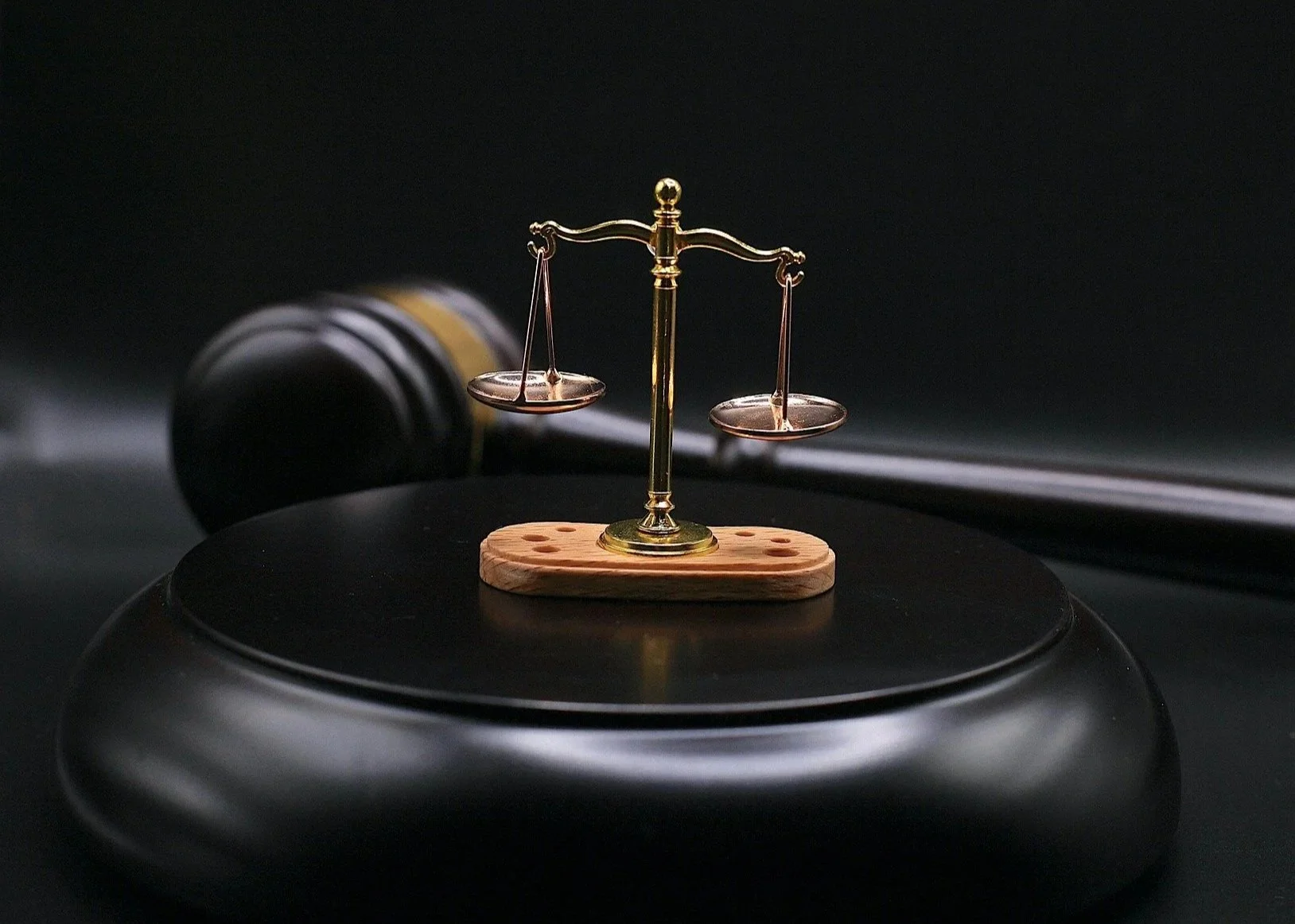 Small golden balance scale on a wooden base, placed on a black surface, with a blurred gavel in the background.