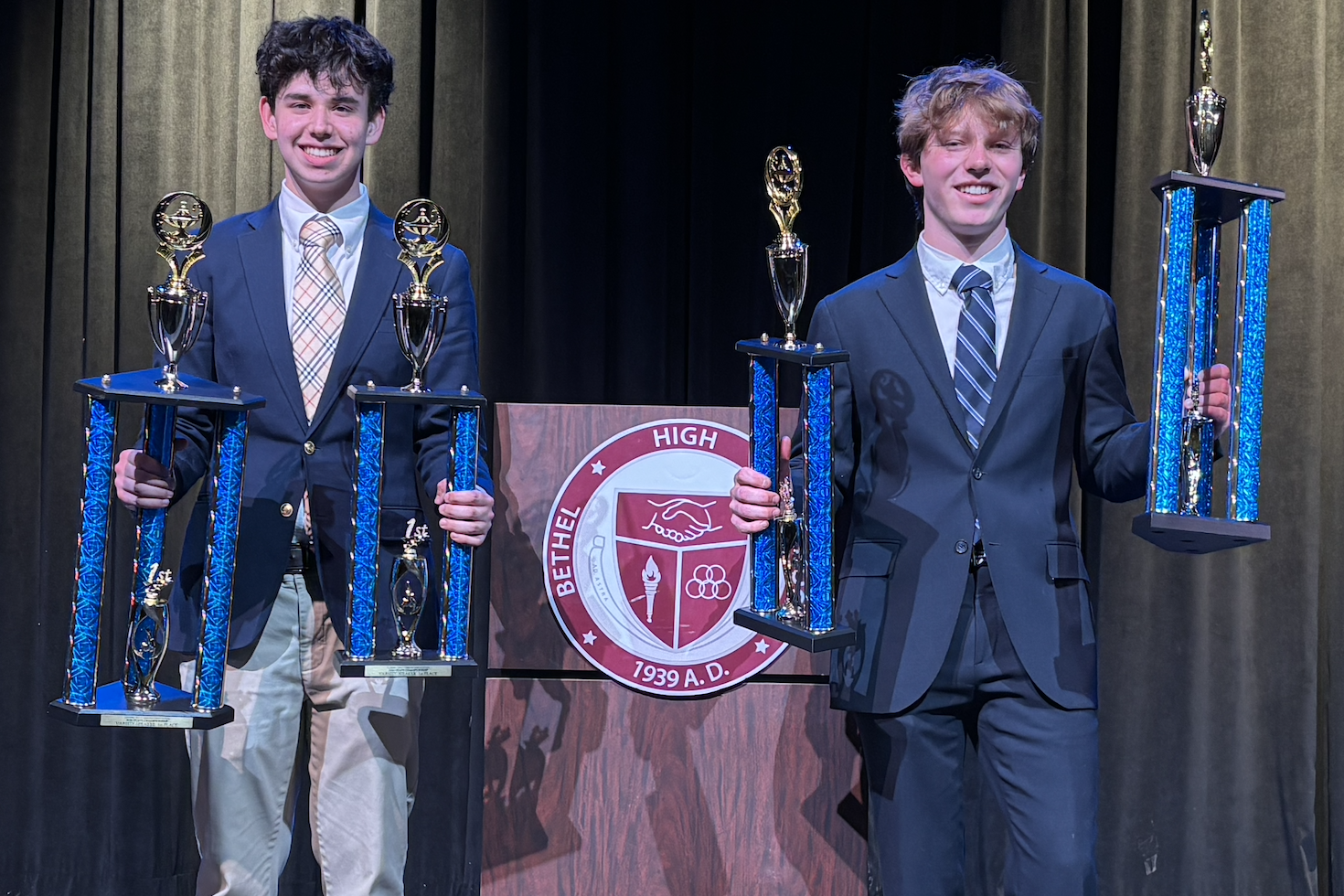 Two young boys in suits holding large trophies, standing on a stage with a school emblem in the background.
