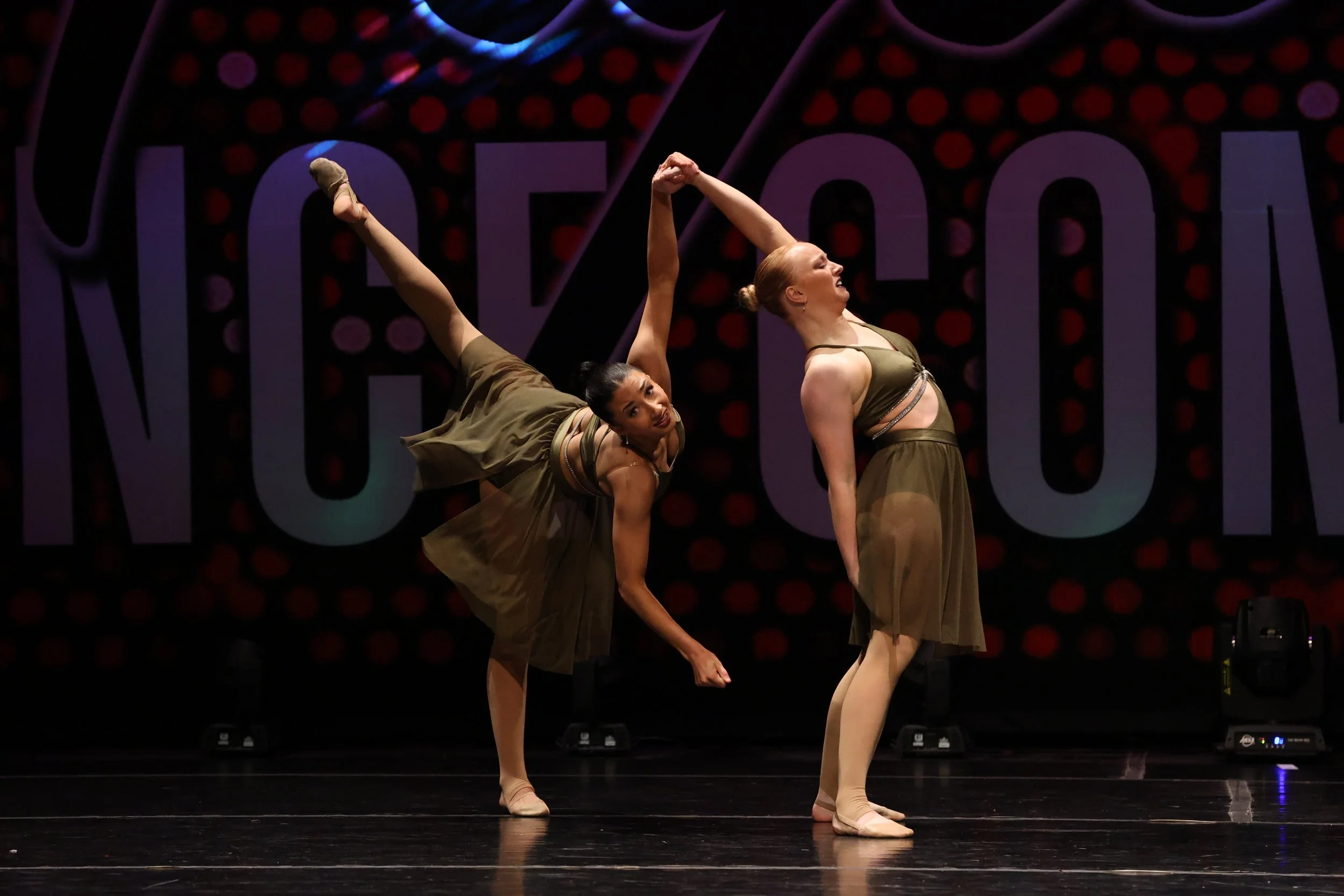 Two ballet dancers perform on stage wearing matching olive green costumes. One dancer is balancing on one leg while the other supports her by holding her hand. The background features a dark backdrop with large purple and red lettering.