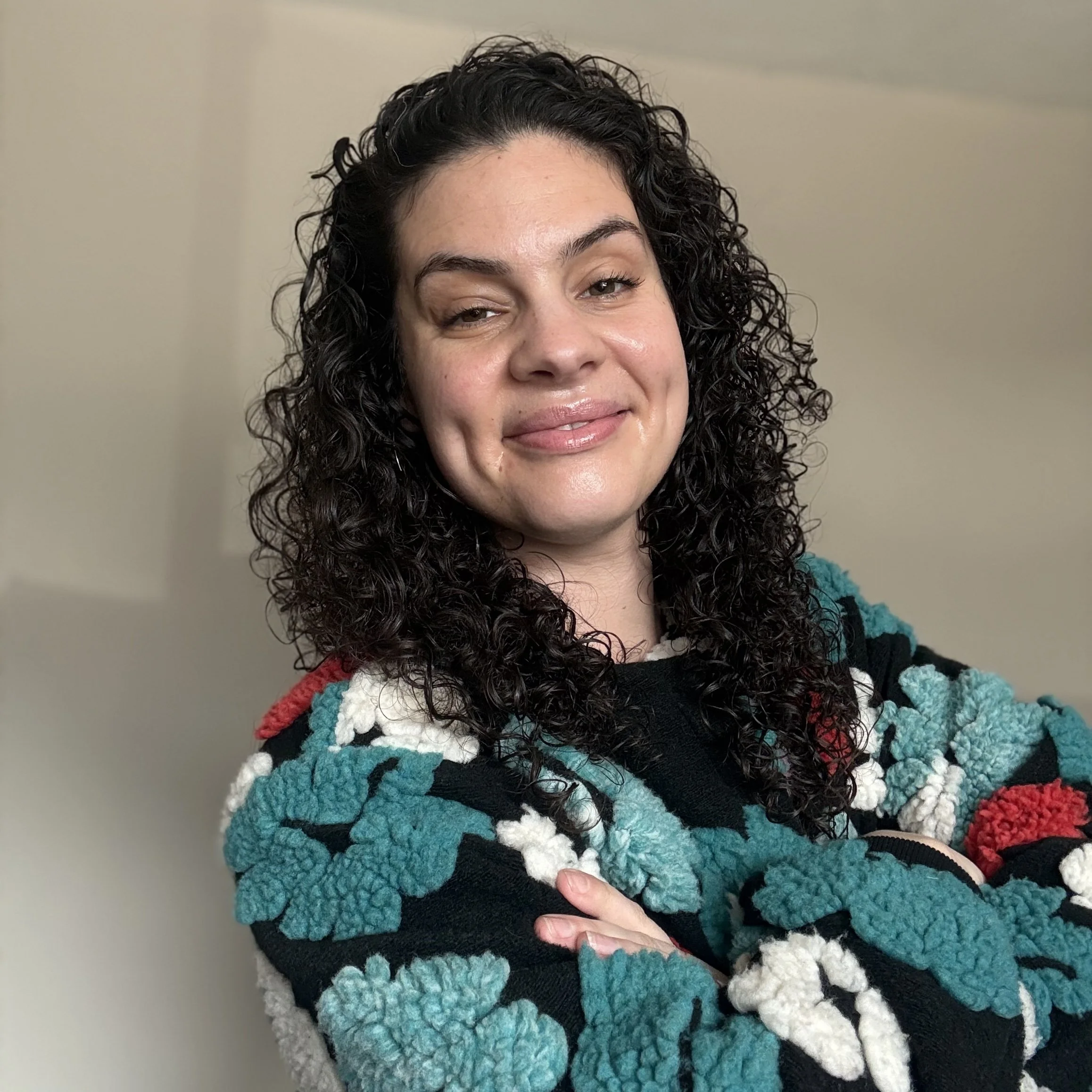 A woman with dark curly hair smiling and crossing her arms, wearing a patterned sweater with teal, black, white, red, and beige colors.
