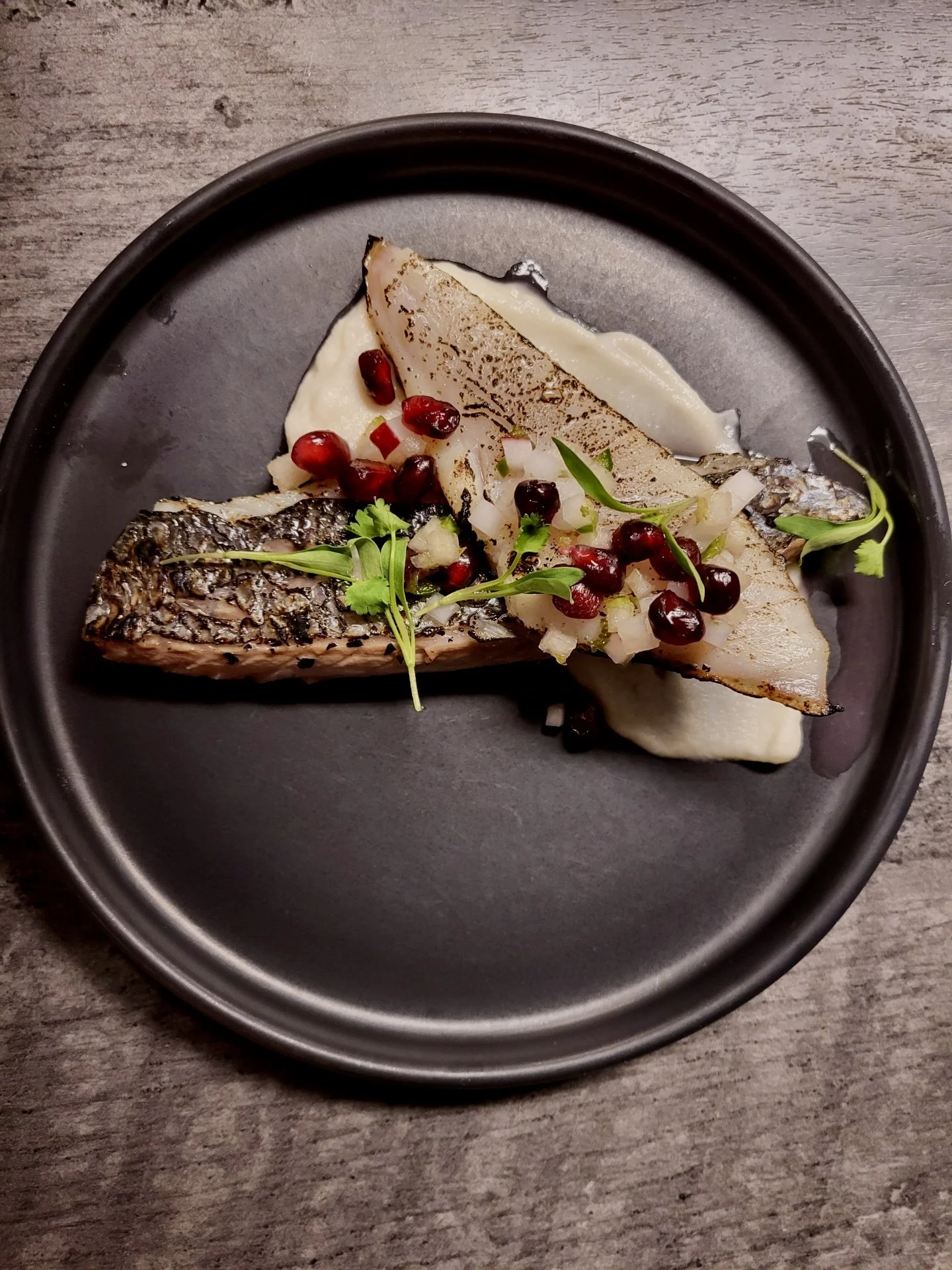 A plated dish with grilled fish, pomegranate seeds, chopped onions, and microgreens, served on a black plate.