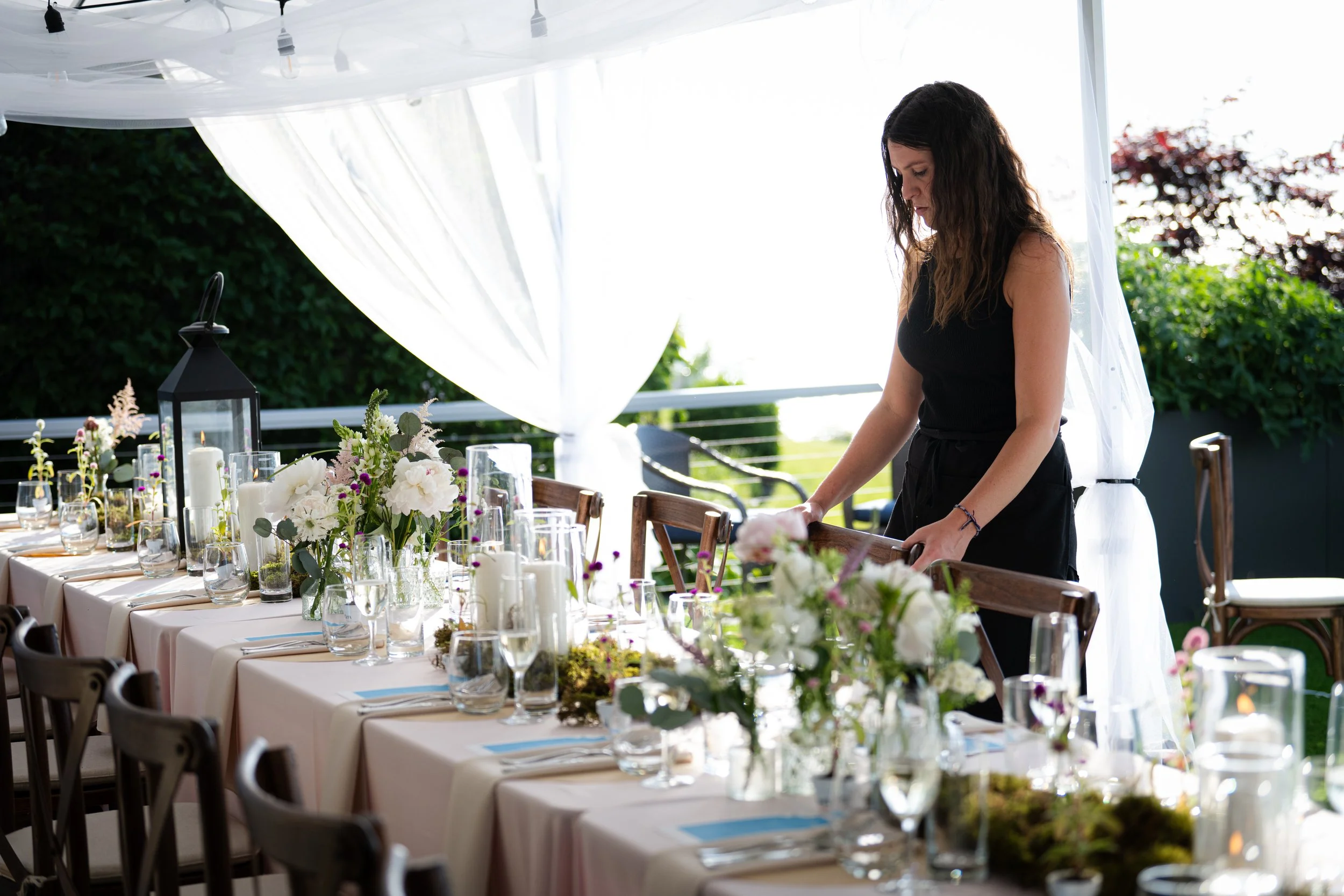 A woman setting up a beautifully decorated outdoor dining table with floral centerpieces, candles, and glassware under a white canopy on a sunny day.