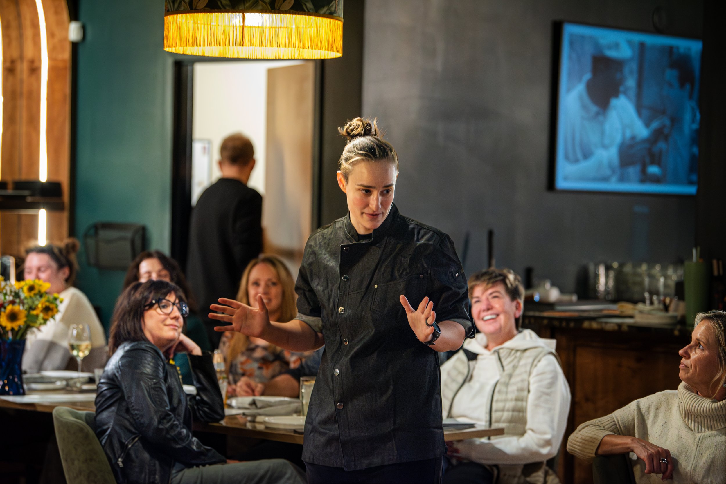 A woman in a black chef's jacket is speaking or performing in front of a group of seated people in a restaurant or event space. The audience is smiling and engaged. The setting has warm lighting, with a large yellow hanging lamp and a television screen on the wall displaying a black and white image.