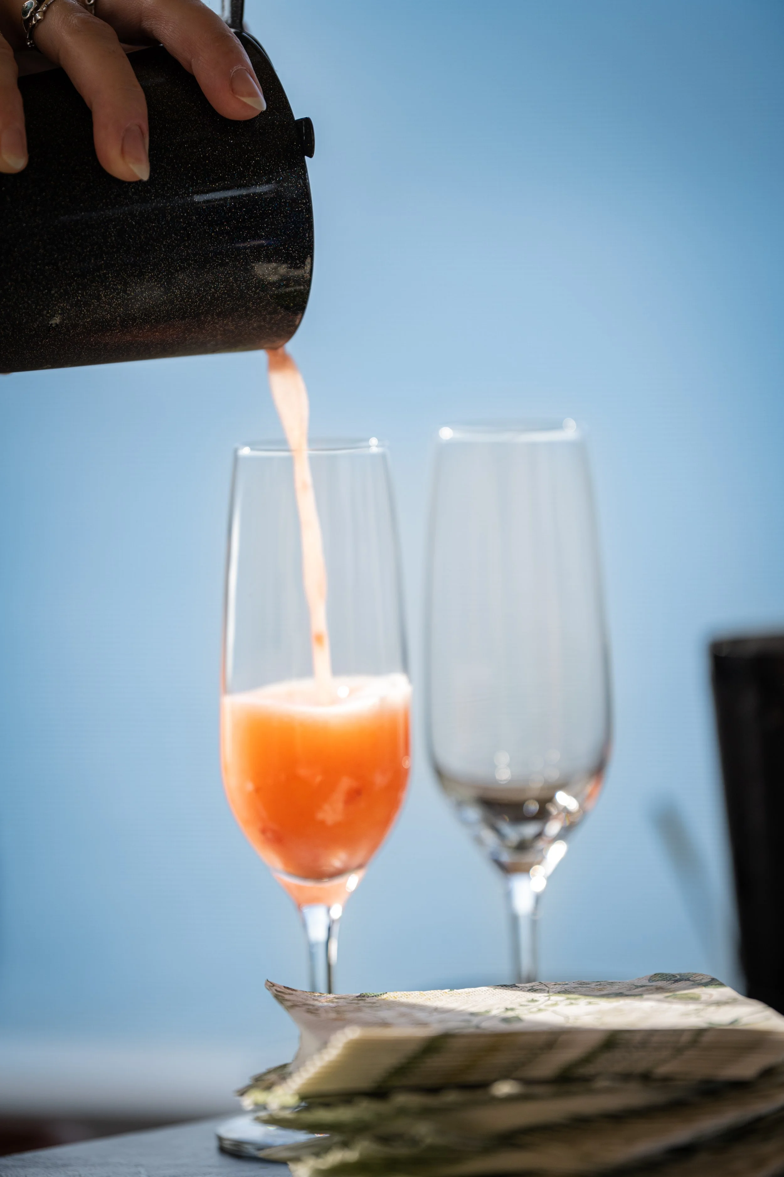 A hand pouring a pinkish-orange beverage from a black sparkly thermos into a tall champagne flute. There is an empty champagne flute next to it and a stack of paper napkins underneath. The background is a light blue color.