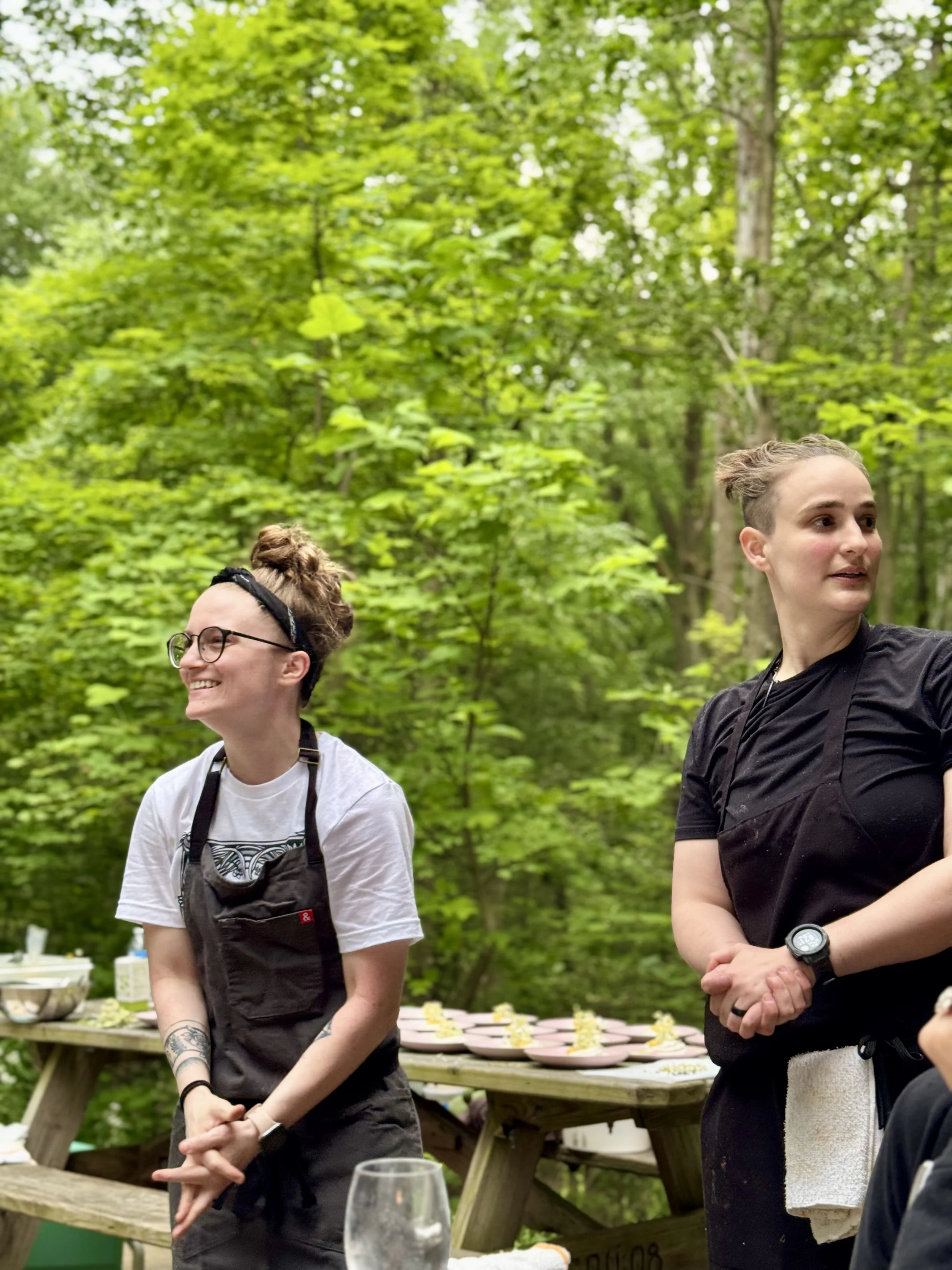 Two women standing outdoors among green trees, smiling and engaged in conversation, with tables and plates in the background, suggesting a social gathering or outdoor event.