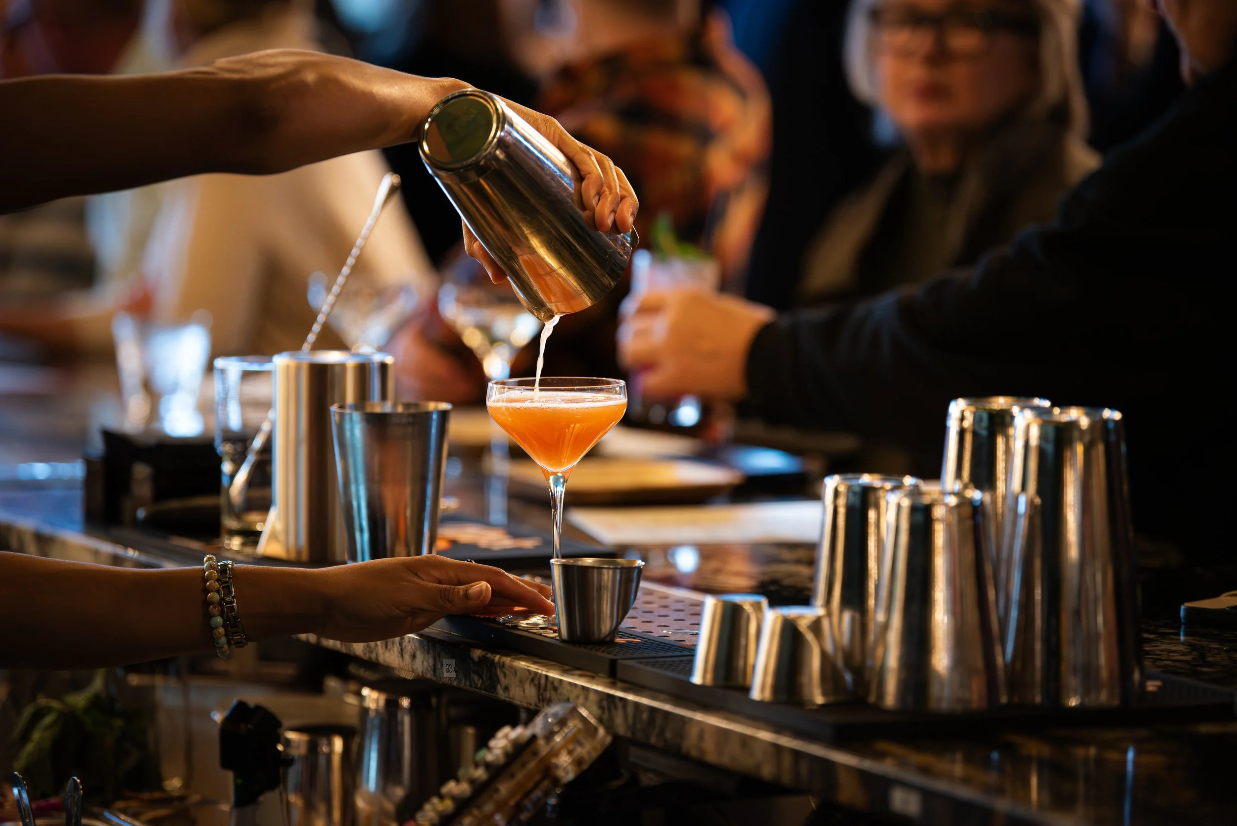 A bartender pouring a cocktail into a glass at a bar with blurred patrons in the background.