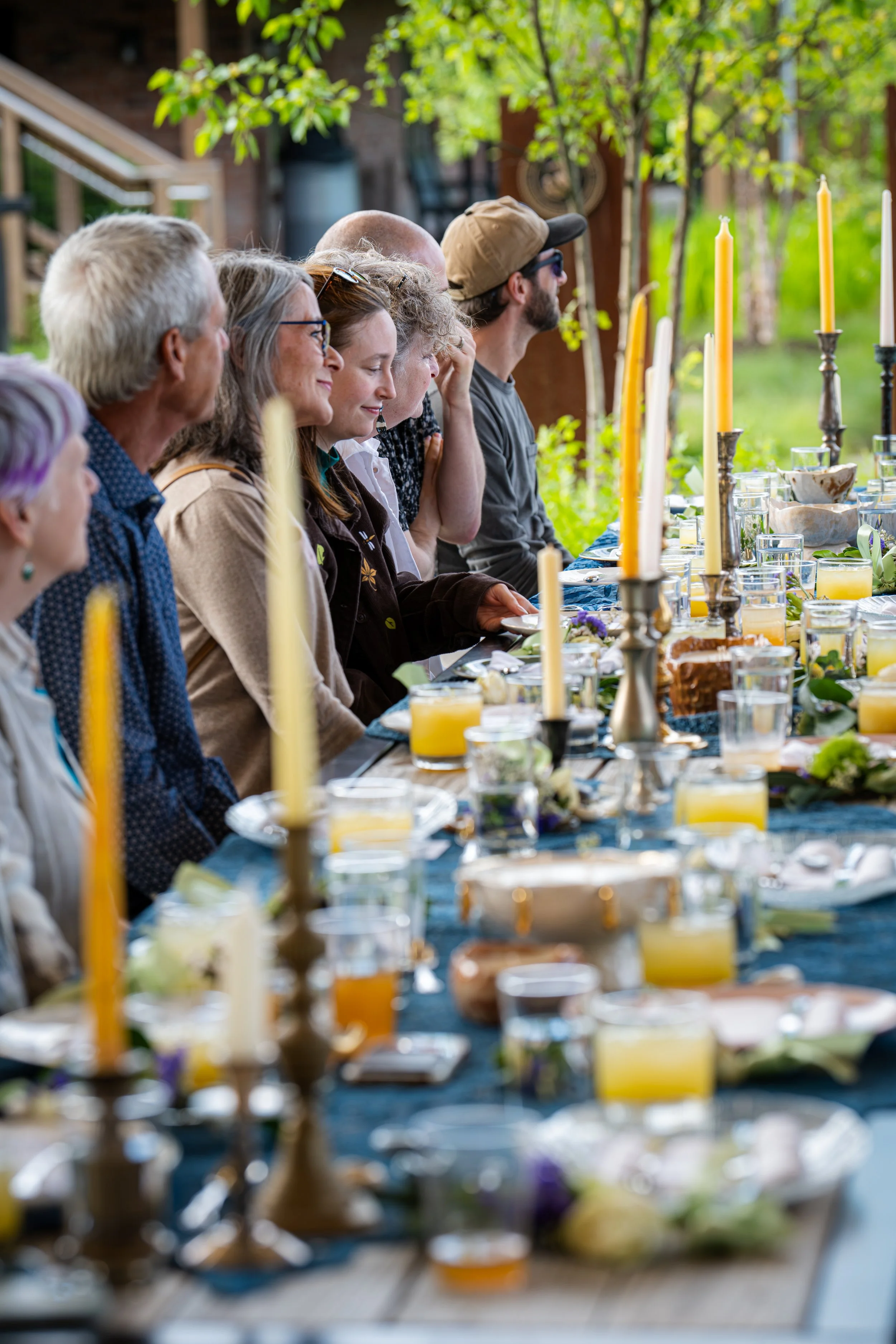 A group of people sitting around a dining table outdoors with greenery in the background. The table is decorated with tall yellow candles, glassware, and floral arrangements.