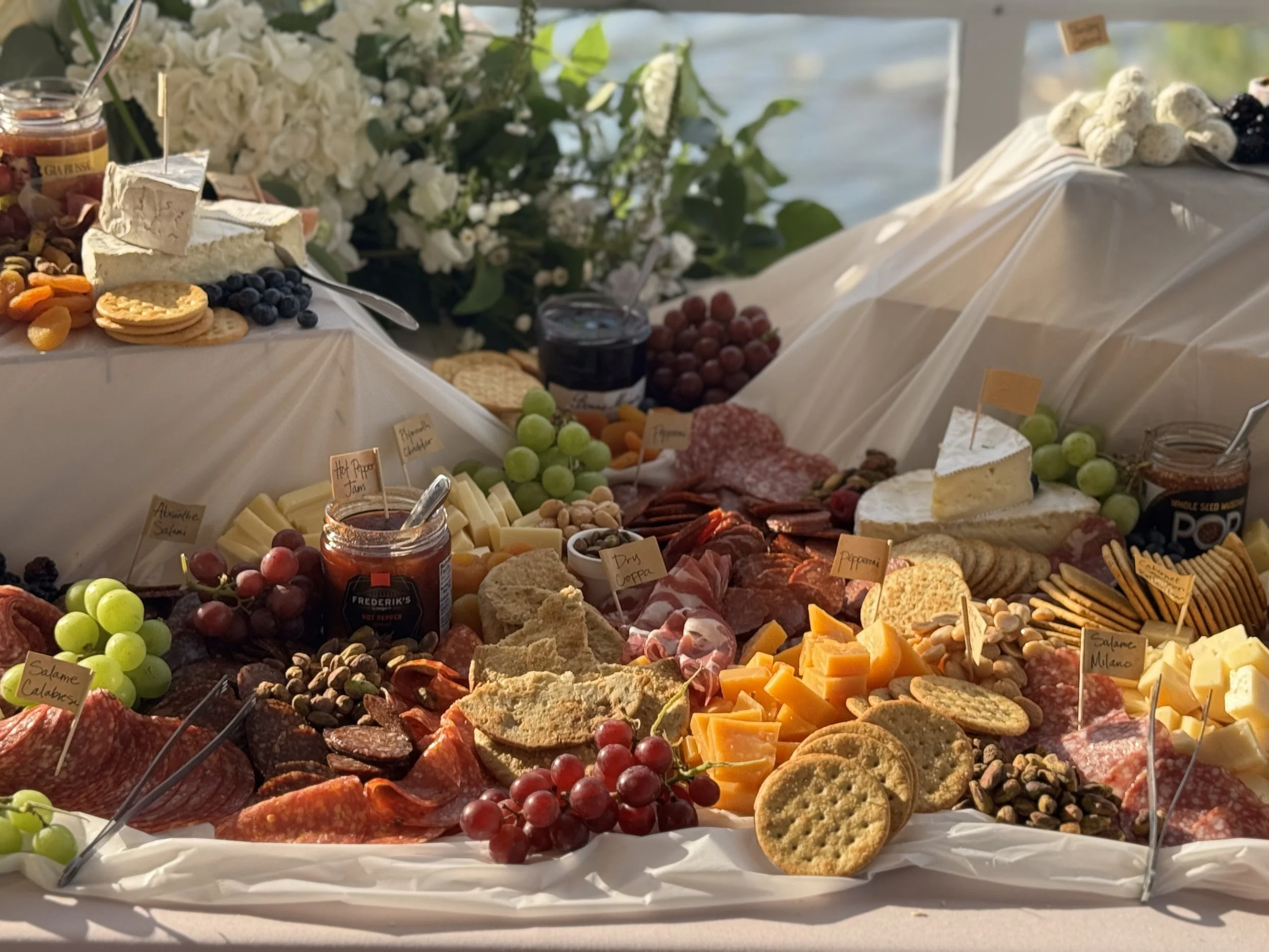 A cheese and charcuterie board with grapes, crackers, jams, and various cheeses, with a floral centerpiece in the background.