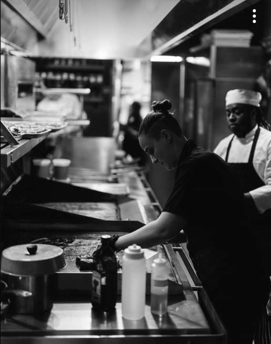 Chef and kitchen staff preparing food in a professional kitchen with shelves and cookware visible.
