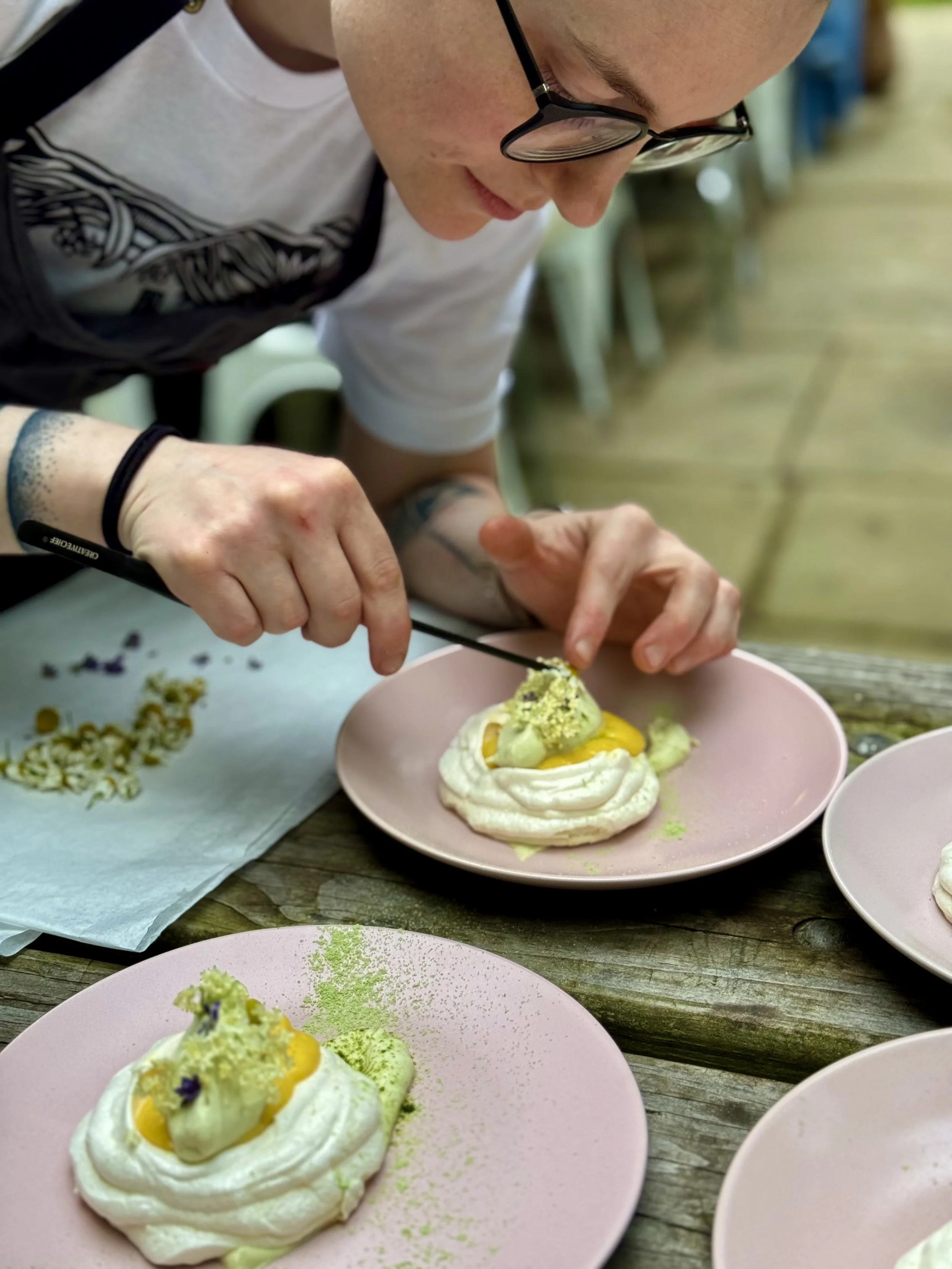 Close-up of a person with glasses decorating a dessert on a pink plate, with other plates of similar desserts on a rustic wooden table.