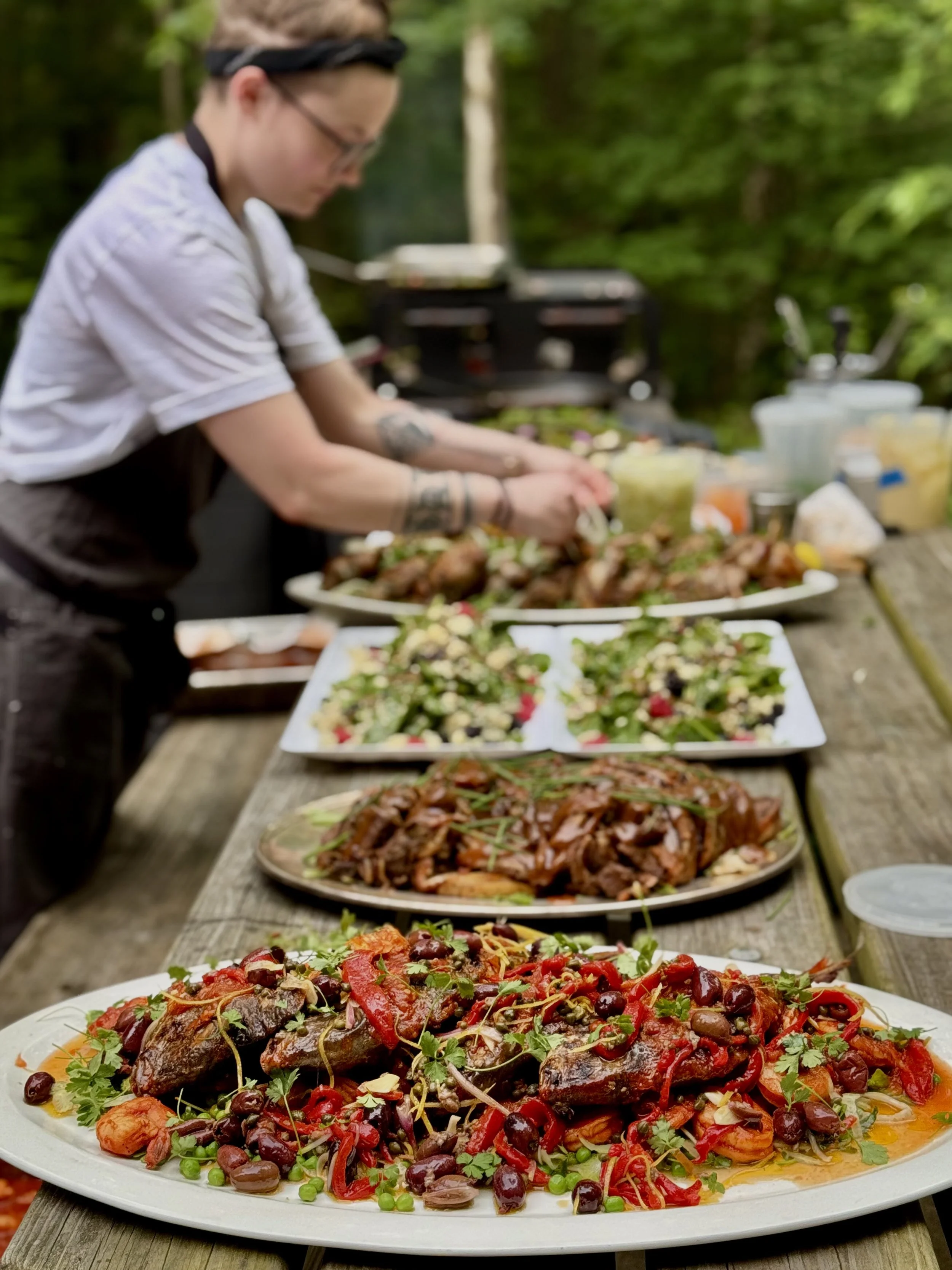 A large plate of seafood with herbs and vegetables front and center, with several other dishes and a blurred outdoor setting in the background.