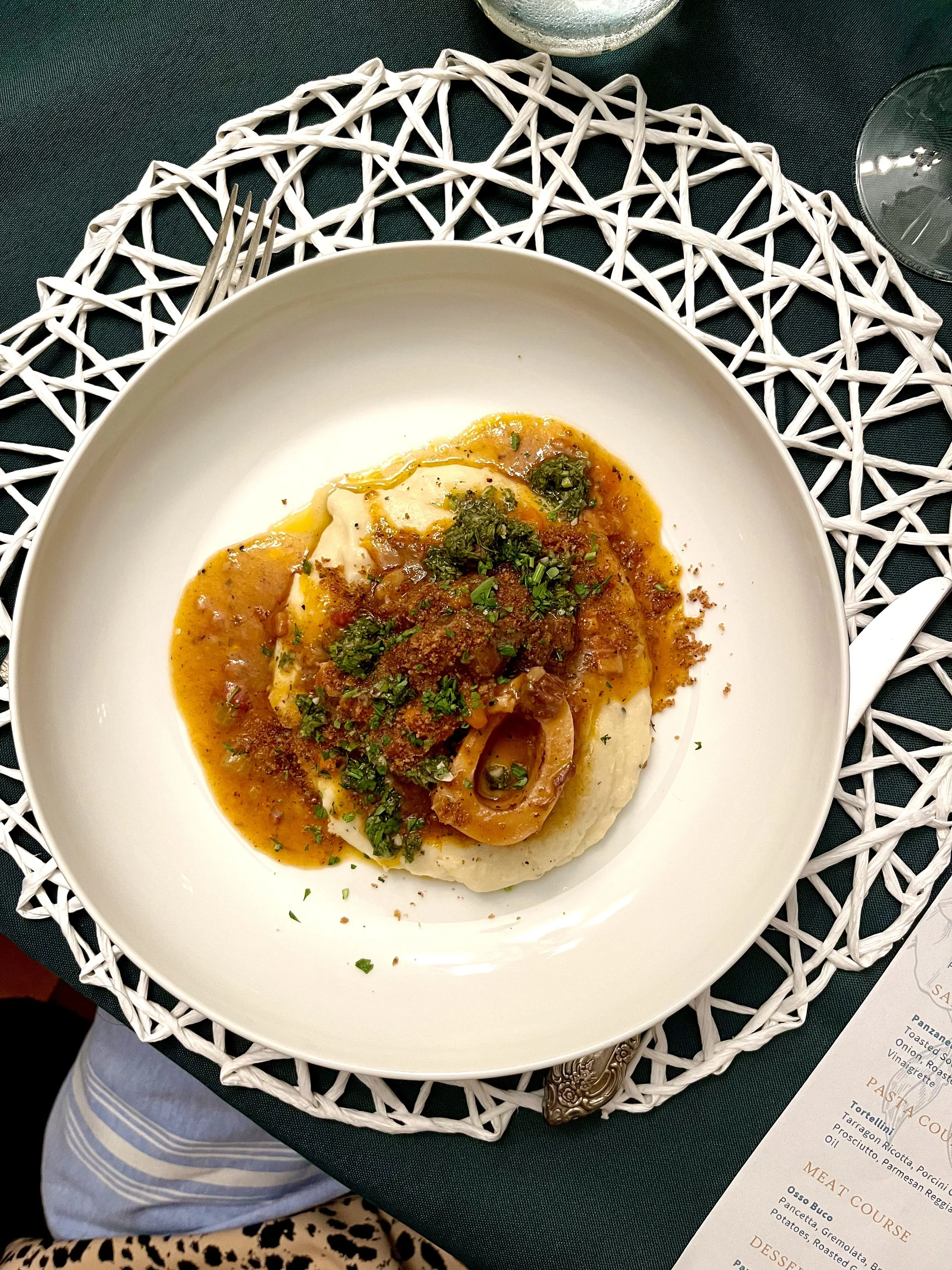 Ossobucco with polenta topped with gravy, herbs, and sauce, served on a white plate on a decorative placemat, with a glass of water, a fork, and a restaurant menu nearby on a dark green tablecloth.