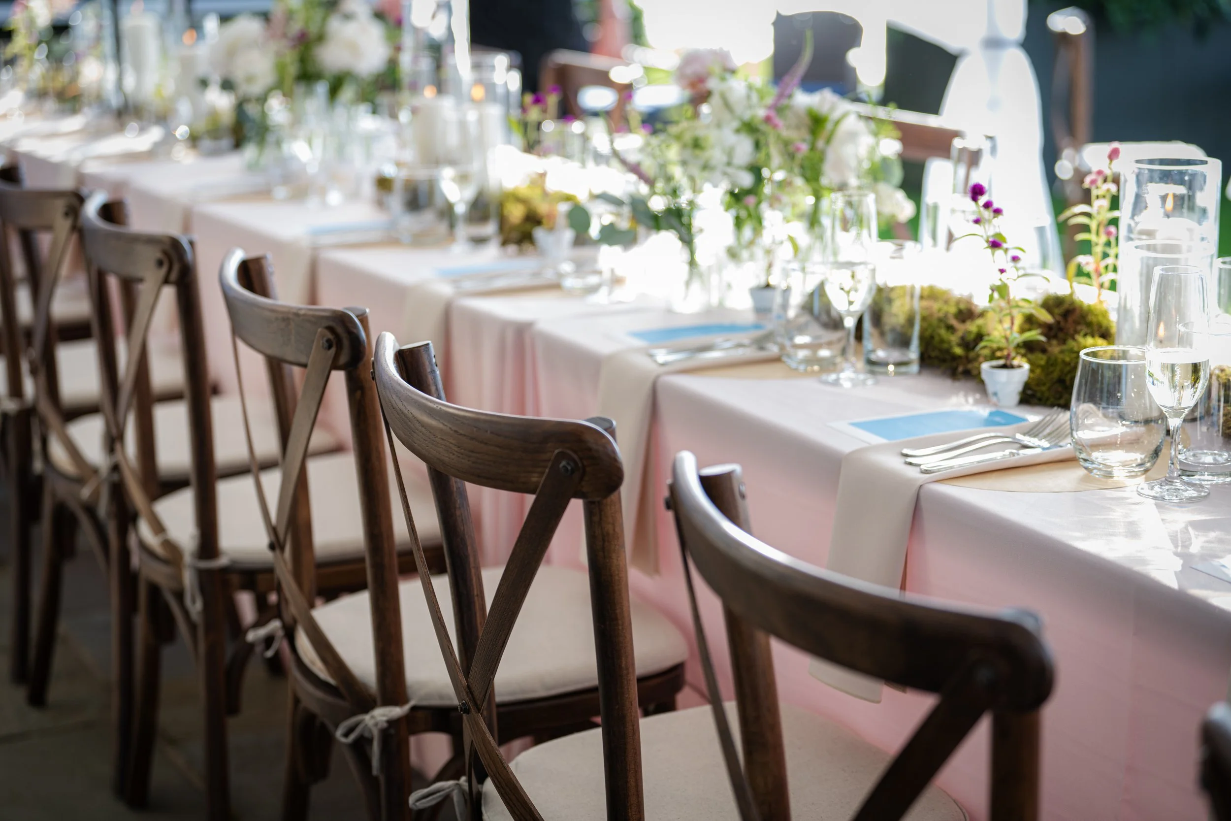 A long dining table set for a formal event with white tablecloths, glassware, silverware, and floral centerpieces. Wooden chairs surround the table, and natural light illuminates the scene.