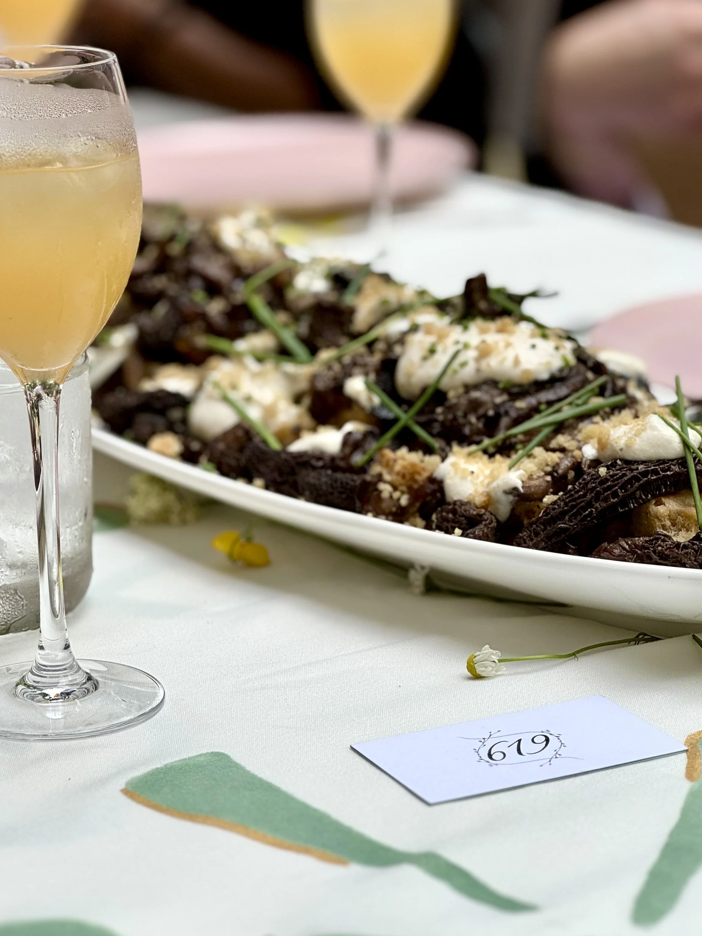 A table setting with a glass of white wine, a floral arrangement, and a plate of food with garnishes, with a sign displaying the number 619.
