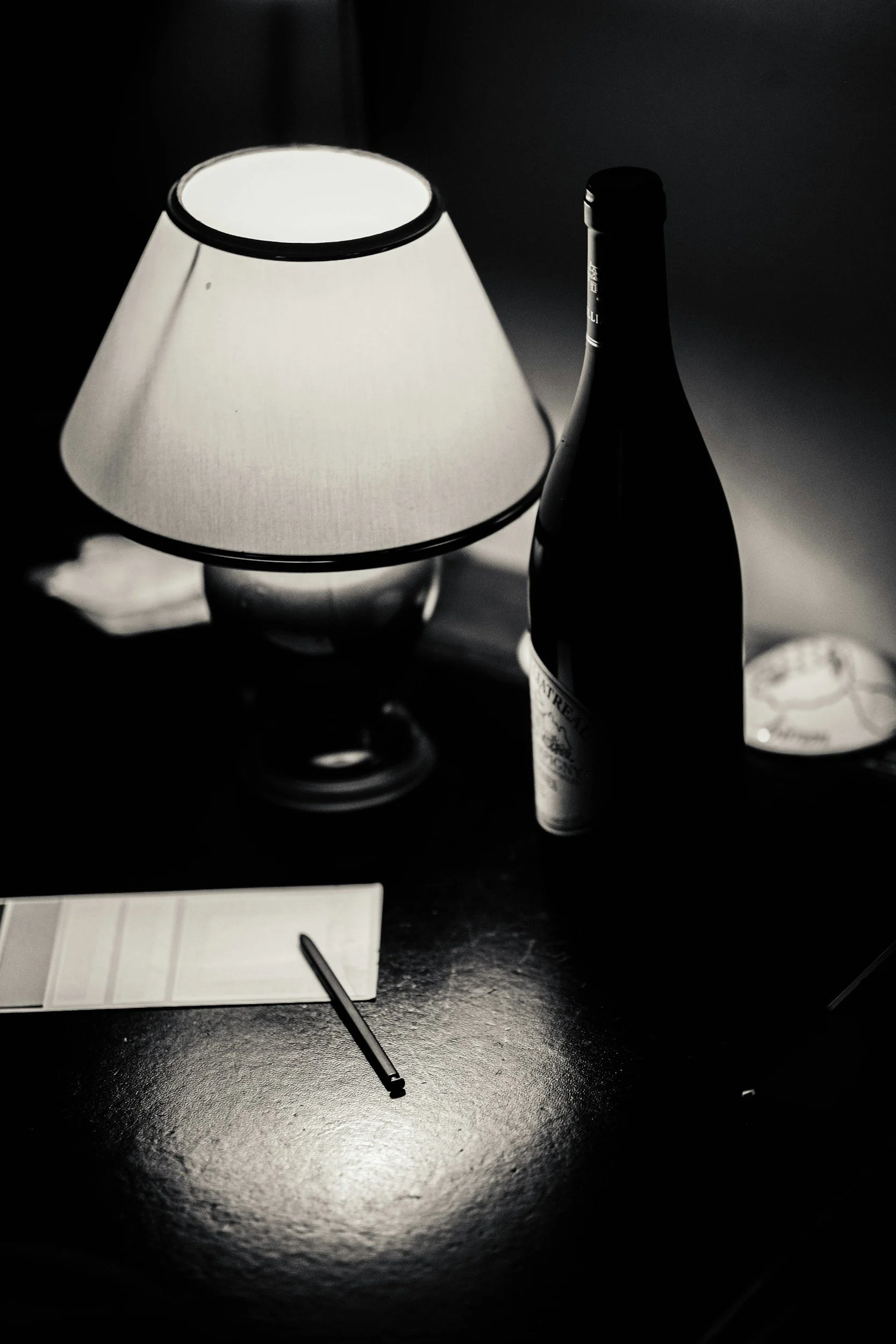 A table with a lamp, a bottle of wine, a notepad, and a pen in black and white.