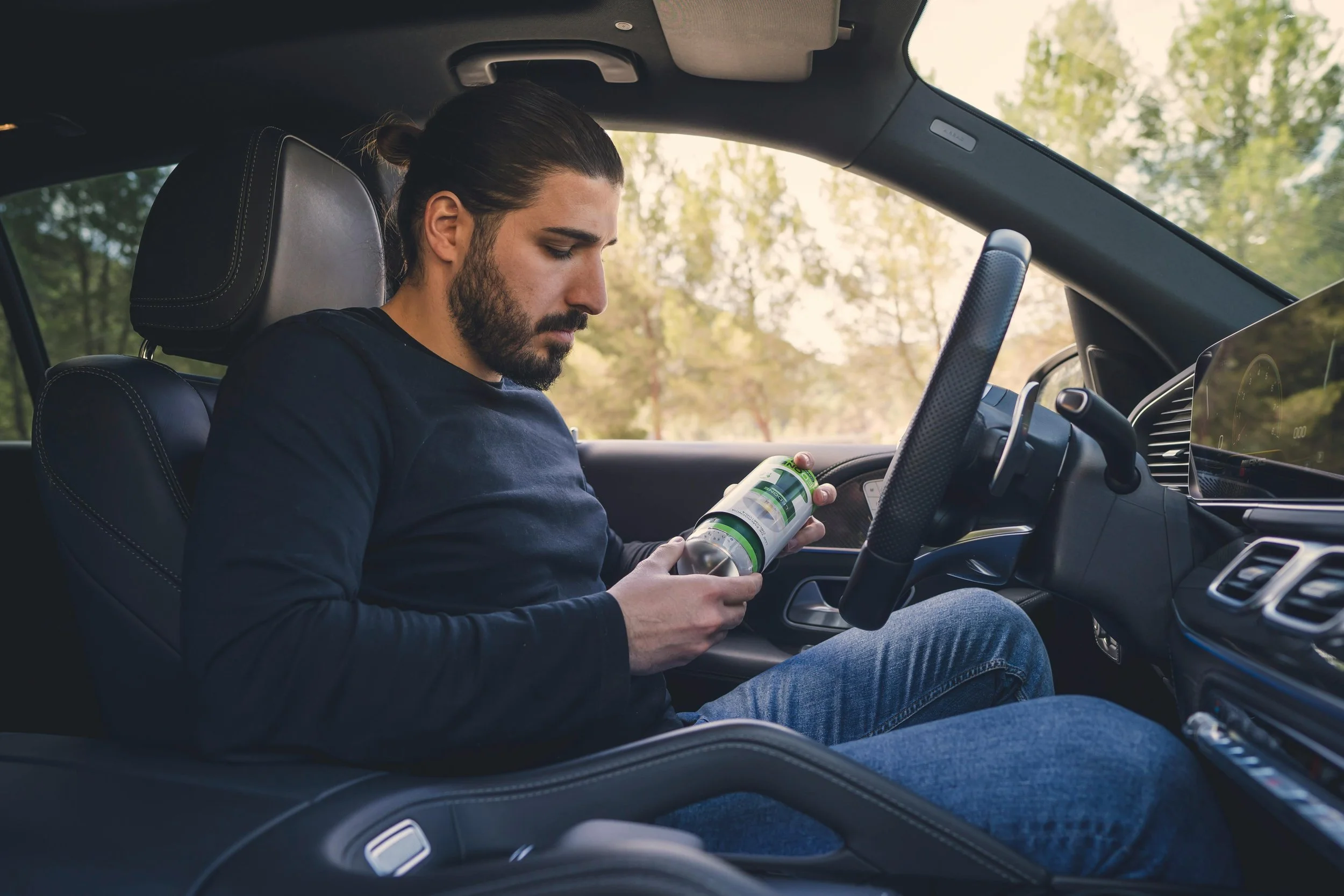 A man with a beard and long hair tied back, sitting in the driver's seat of a car, holding a bottle and looking at his phone, with a background of trees visible through the window.