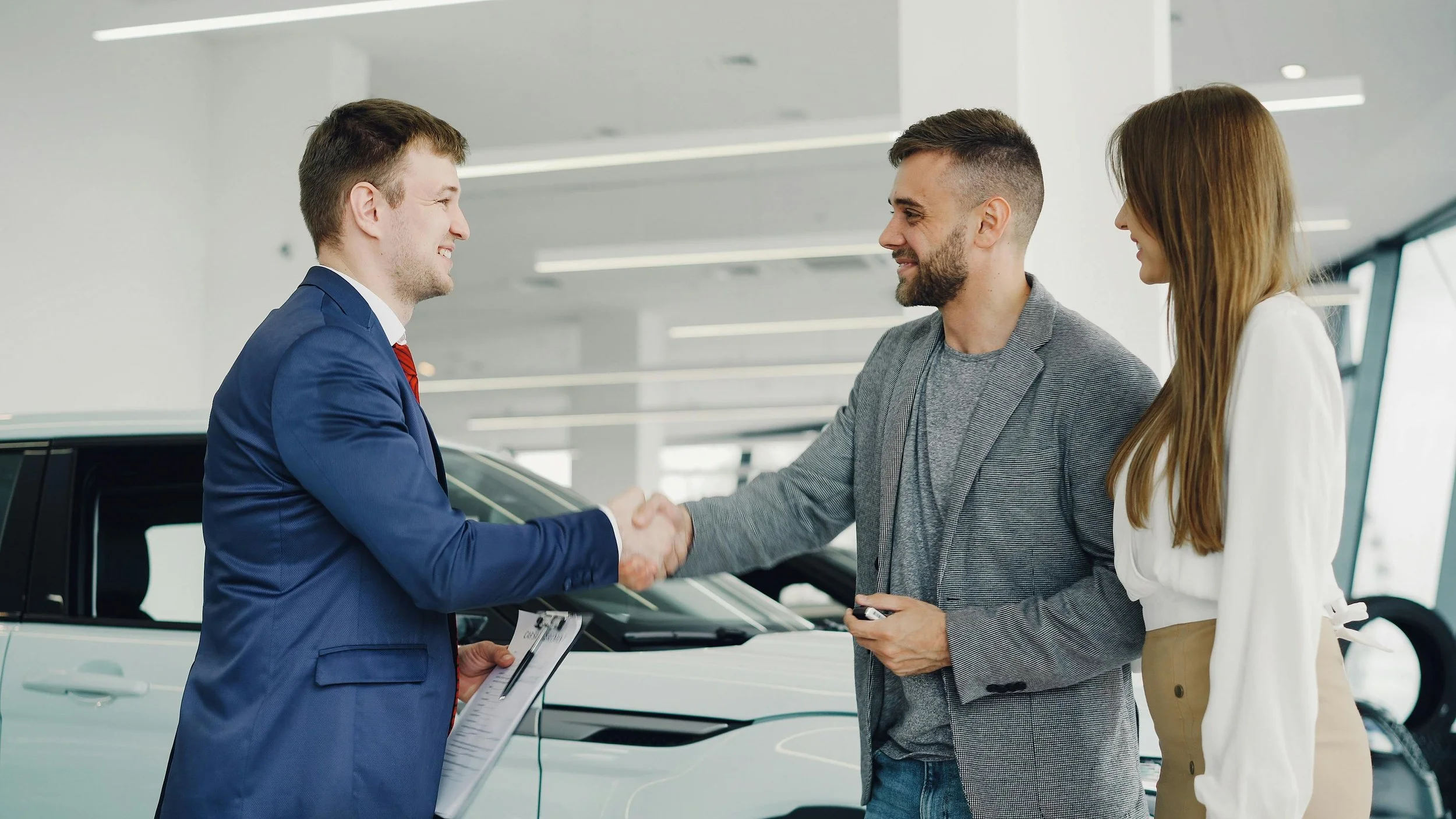 A man in a blue suit shaking hands with another man in a gray blazer while a woman in white stands watching in a car dealership.