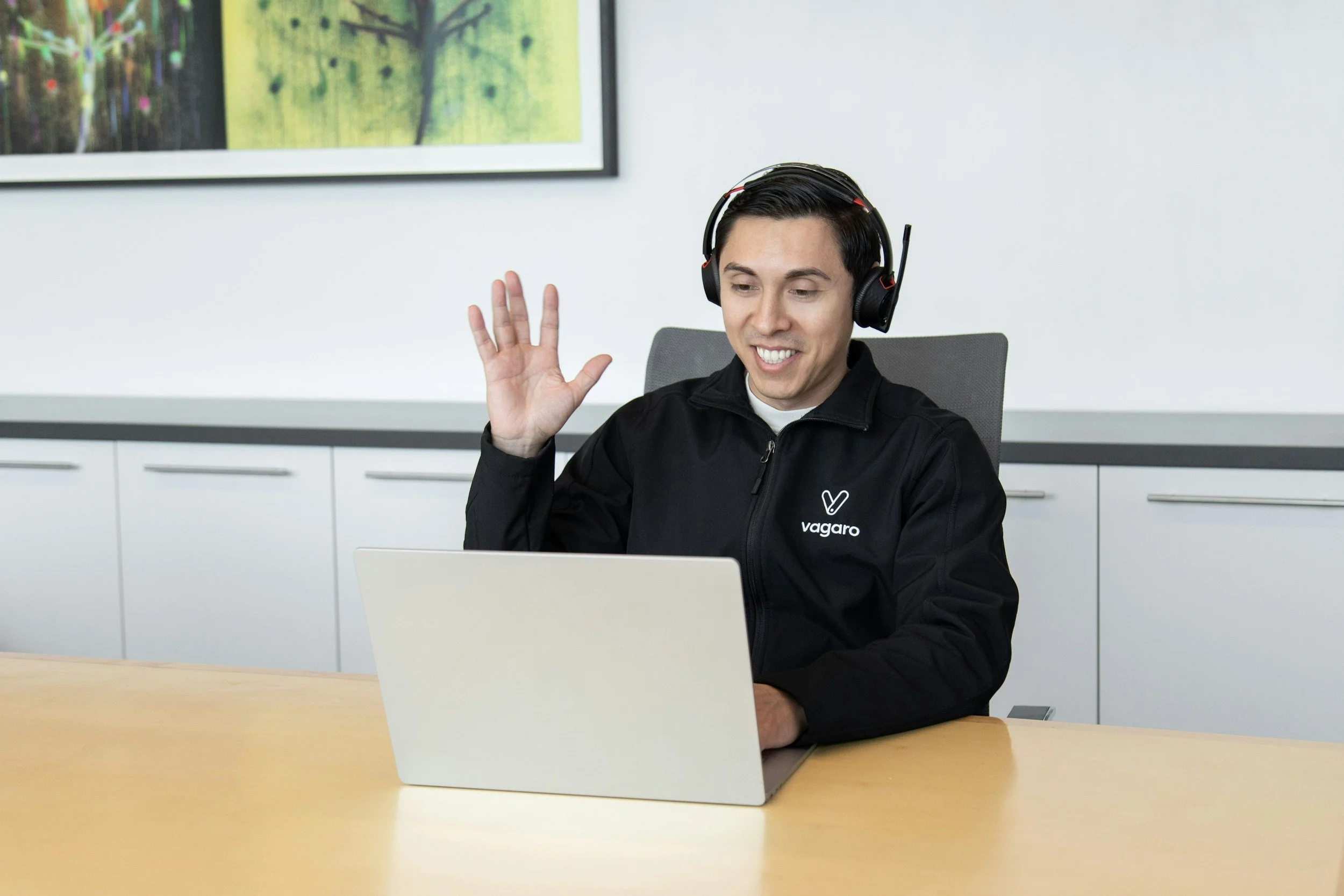 A man with dark hair wearing headphones and a black jacket with a logo sitting at a desk, waving and smiling in front of a laptop, in a modern office setting.