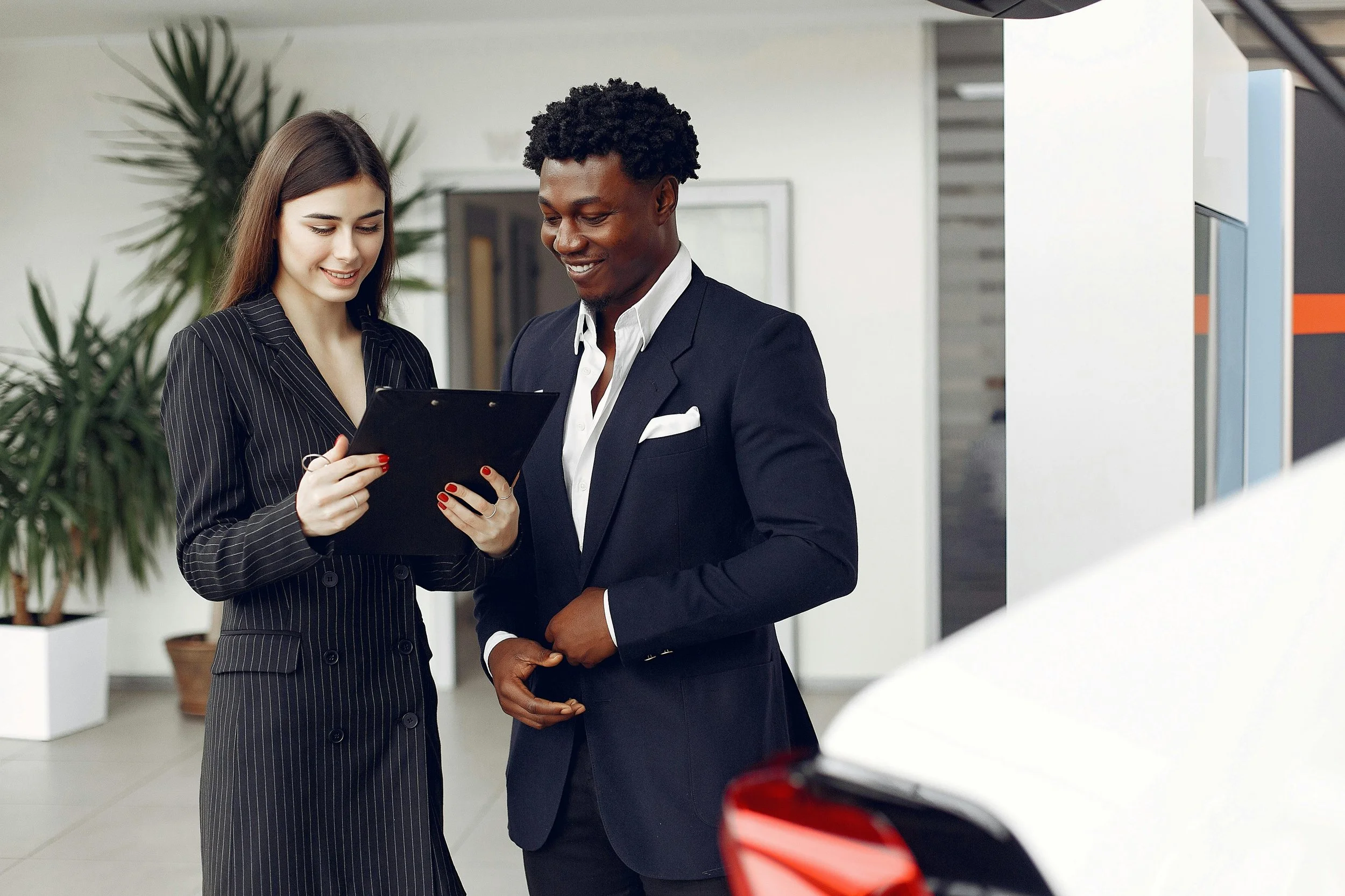 A woman and a man in business attire are standing next to each other, looking at a clipboard the woman is holding. They are smiling and appear to be discussing something. The background includes indoor plants and a white wall.