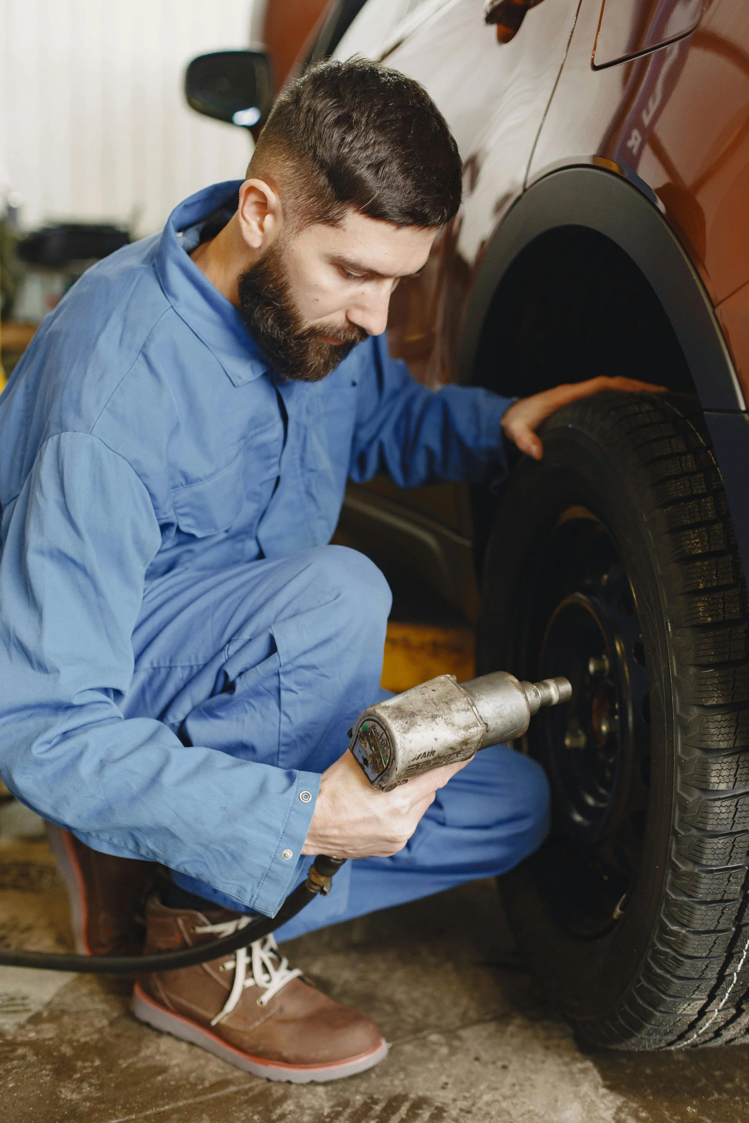 A man in blue overalls working on a car tire using a tool in a garage.