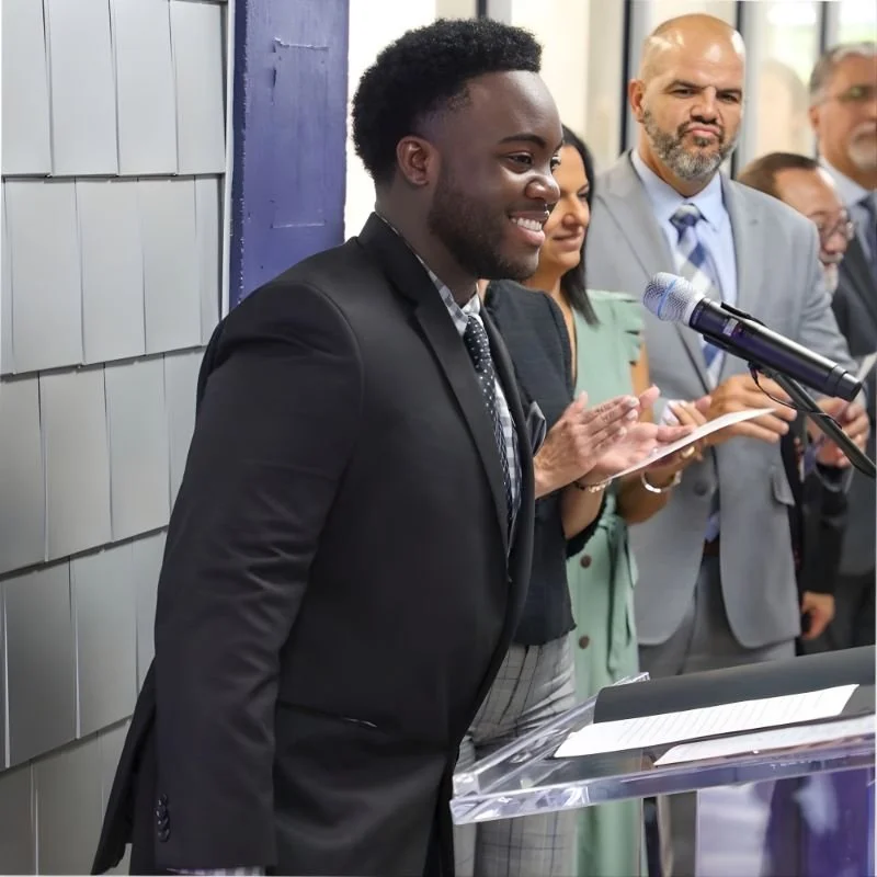 A young man in a black suit speaking at a podium with a microphone, with four other professionally dressed people standing behind him in a formal event or ceremony.