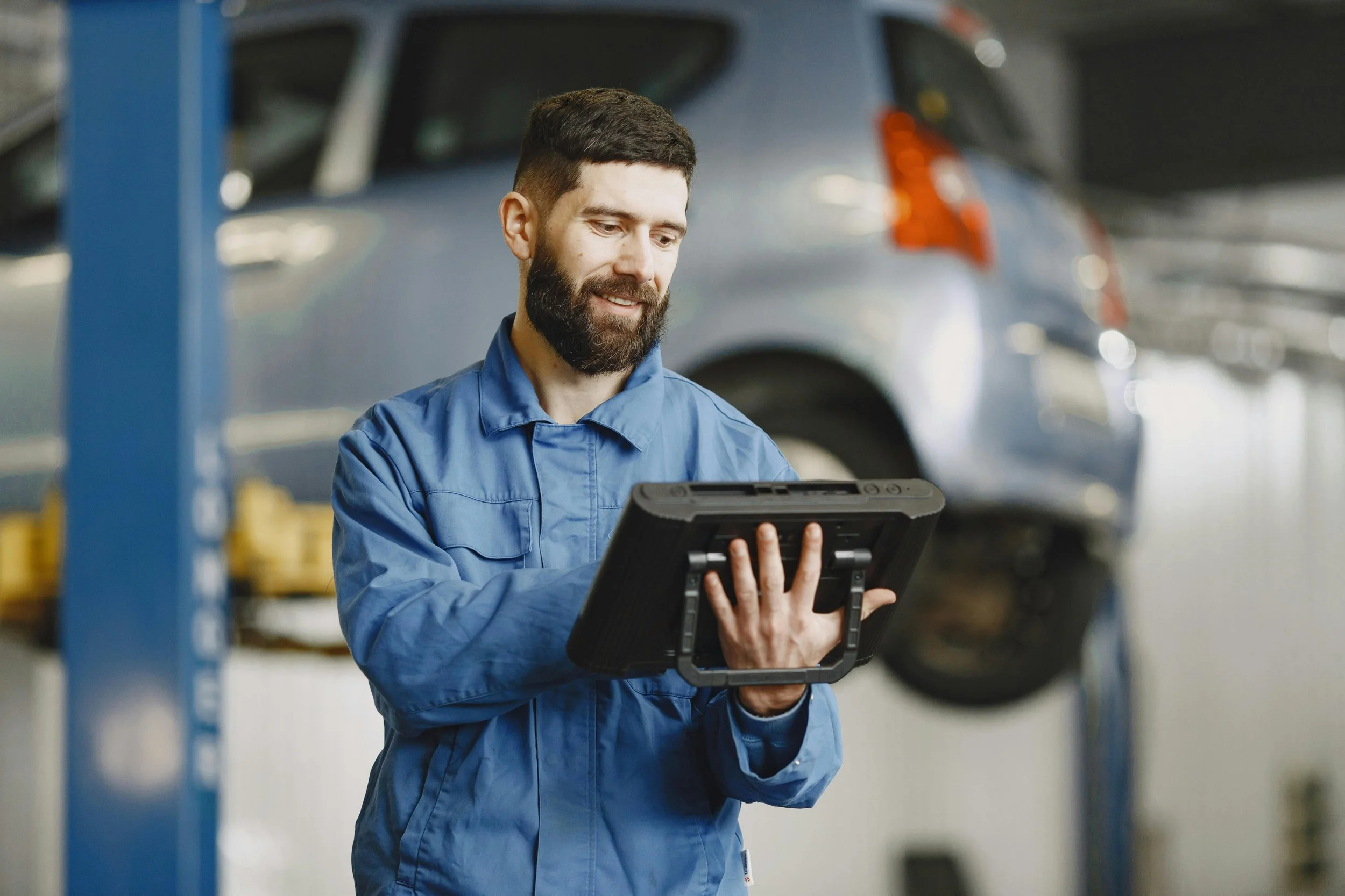 A mechanic in a blue work uniform holding a diagnostic device in an automotive repair shop. A car is lifted on a hydraulic lift in the background.