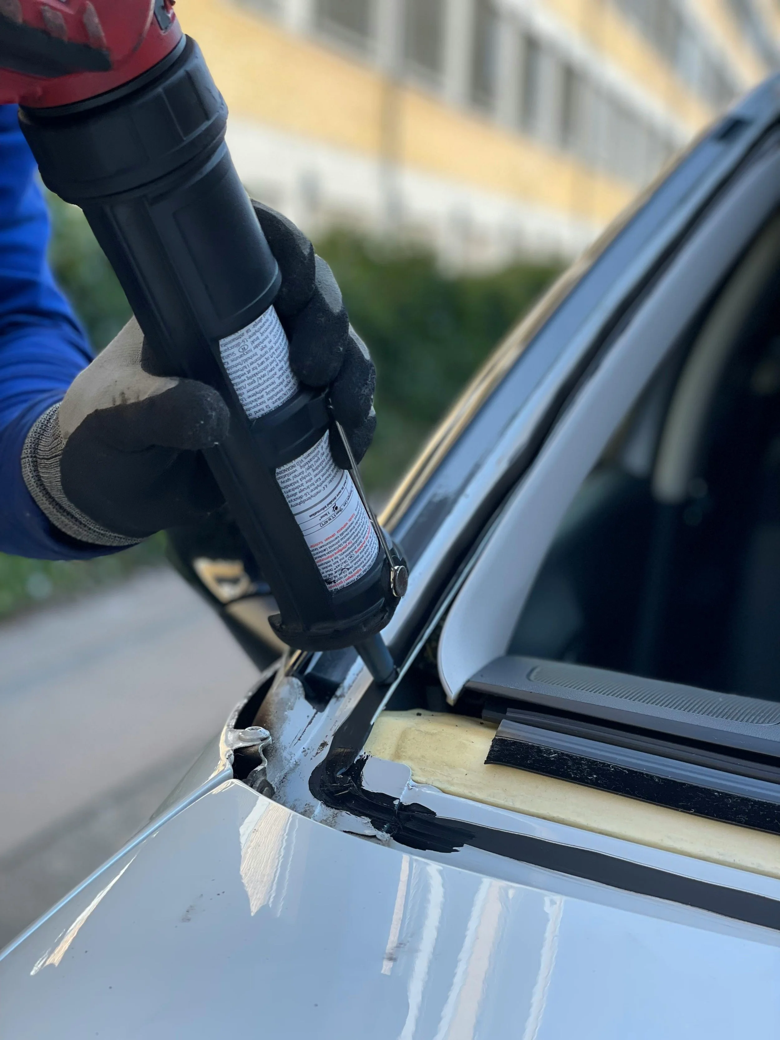A person in black gloves is using a power drill to remove or repair part of a white car, focusing on the area near the dashboard and windshield.