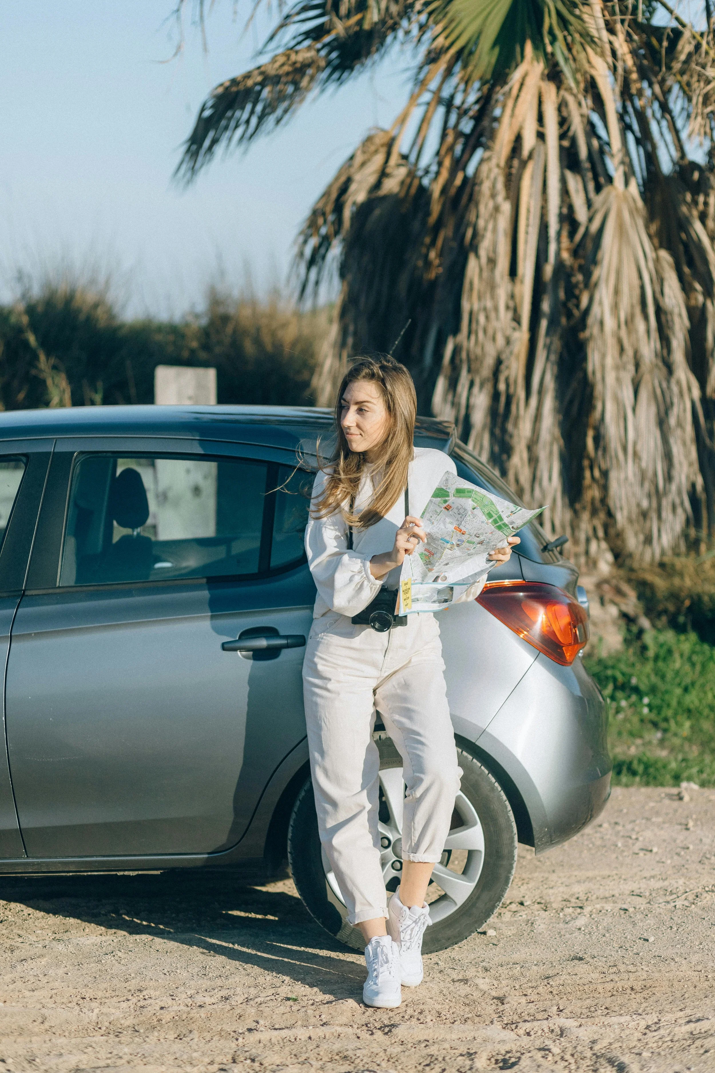 A young woman holding a map while standing next to a silver car on a dirt road with a large palm tree in the background.