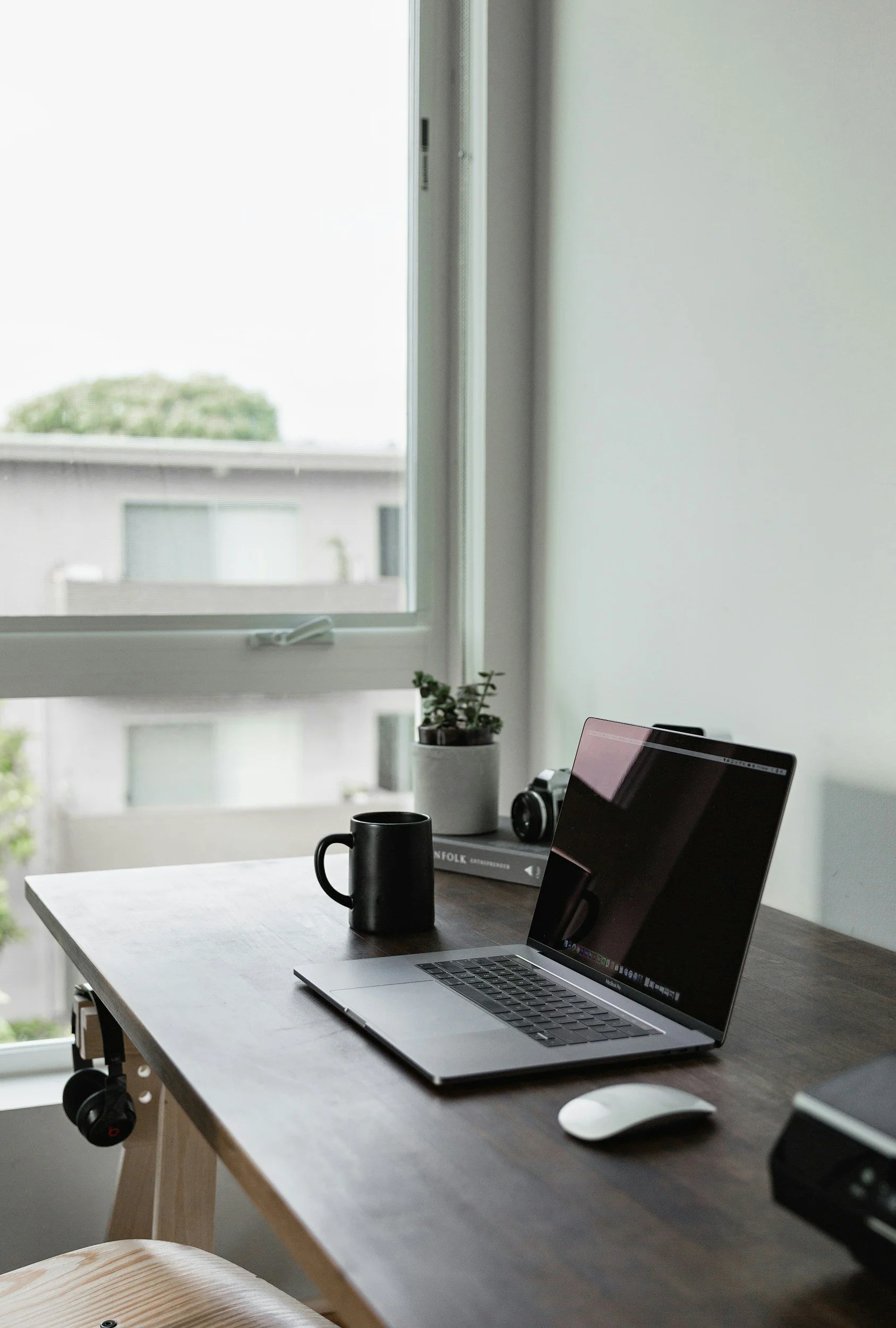Home office desk with a laptop, black mug, white mouse, potted plant, camera, and books near a large window.