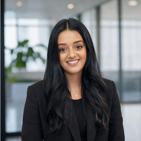 A young woman with long dark hair, wearing a black blazer, smiling in a modern office setting.