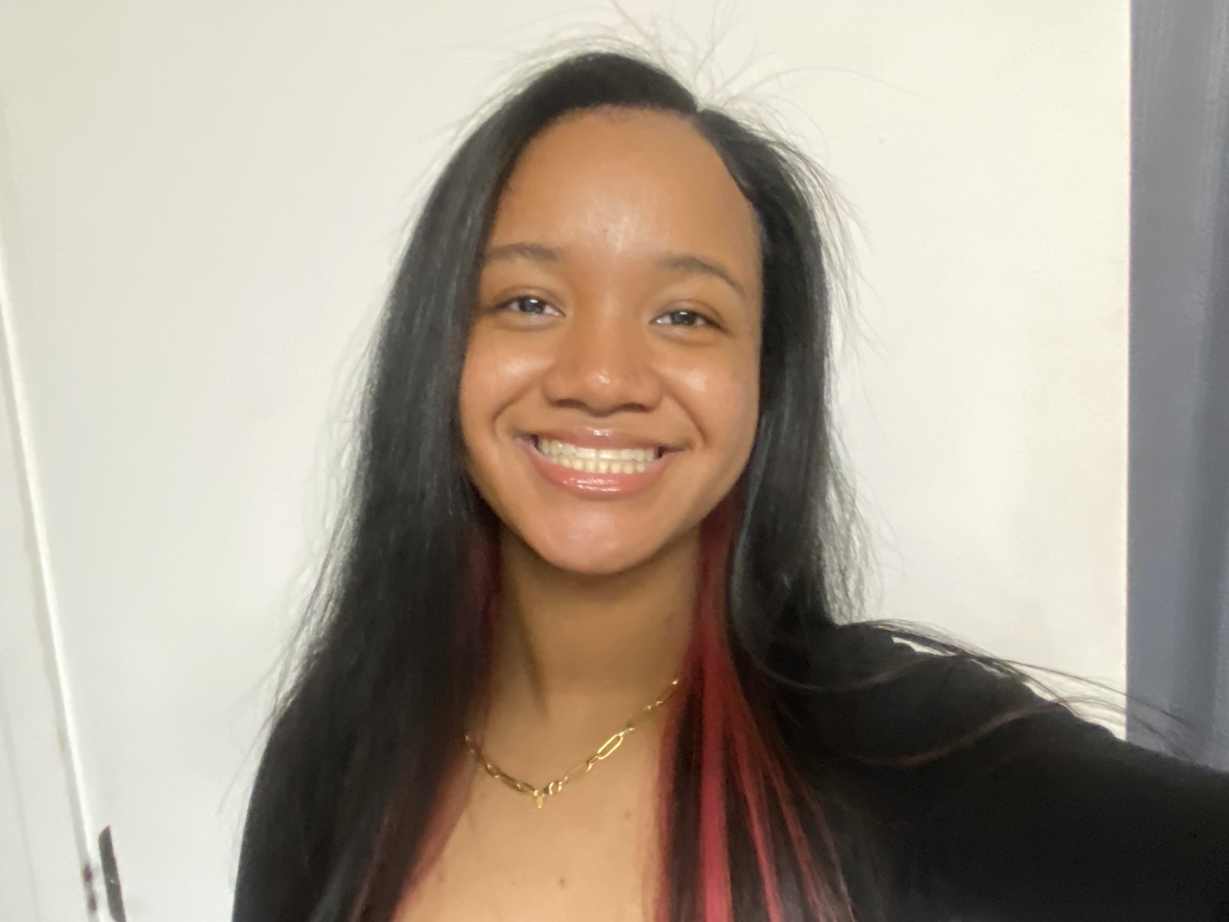 A young woman with long black hair, smiling with braces, wearing a red top and gold necklace, standing against a plain white and gray background.