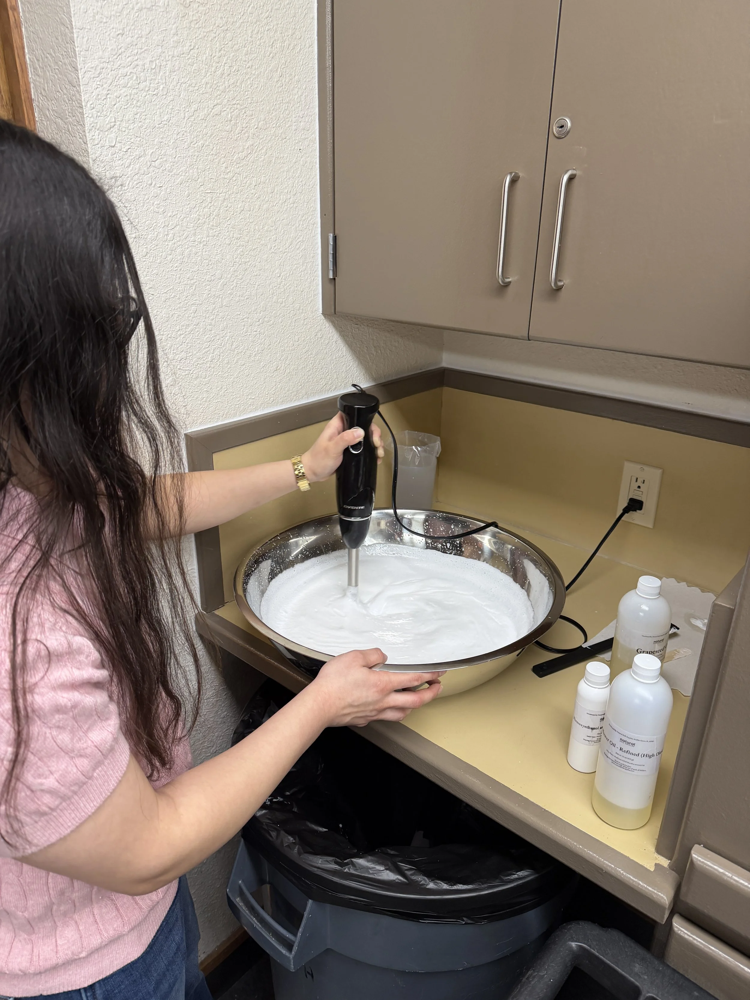 A person wearing a pink sweater and blue jeans holding a black handheld tool with a white foam substance in a stainless steel sink. Several bottles of liquids are on the beige counter nearby. A black trash bin is underneath the sink.